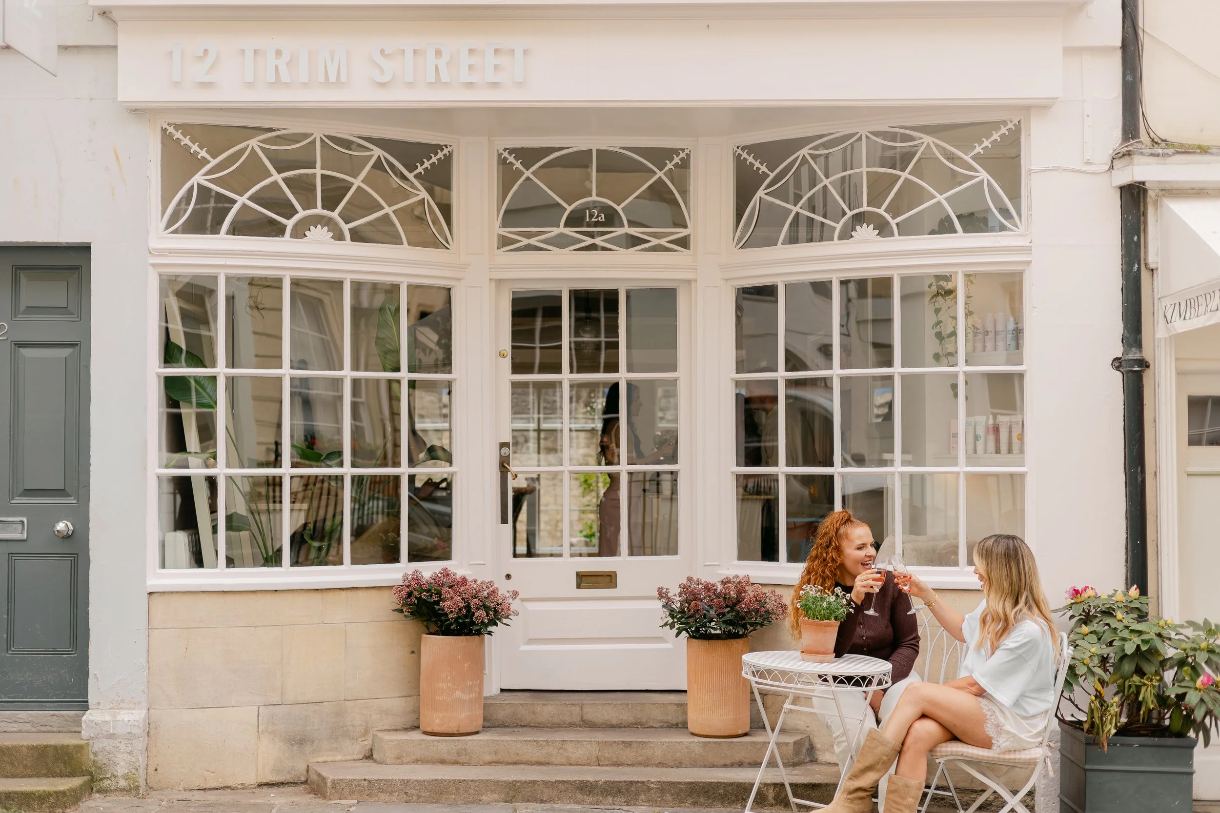 Two women sitting outside a café or restaurant, enjoying drinks and smiling at each other, in front of a storefront with large windows and potted plants. One woman has red hair and the other has blonde hair.