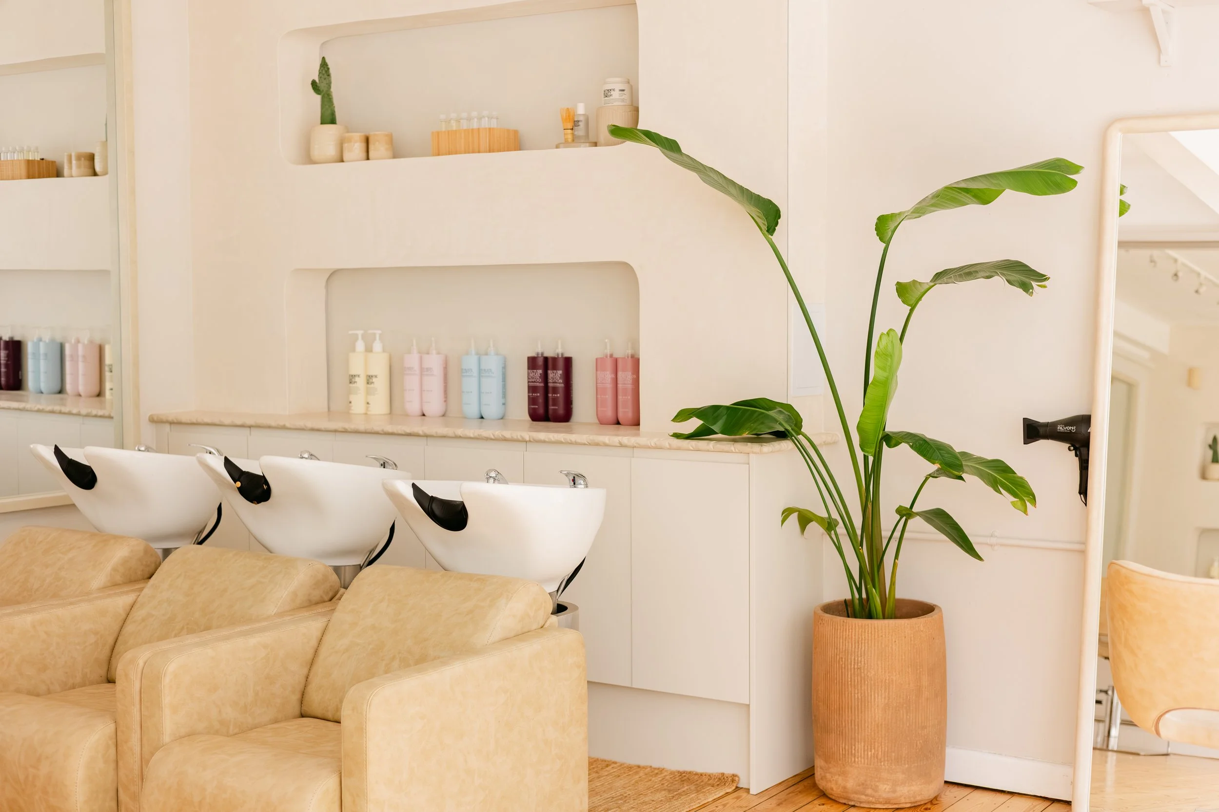 A salon interior with white washing stations and beige sofas, decorated with green plants and shelves filled with various hair products.