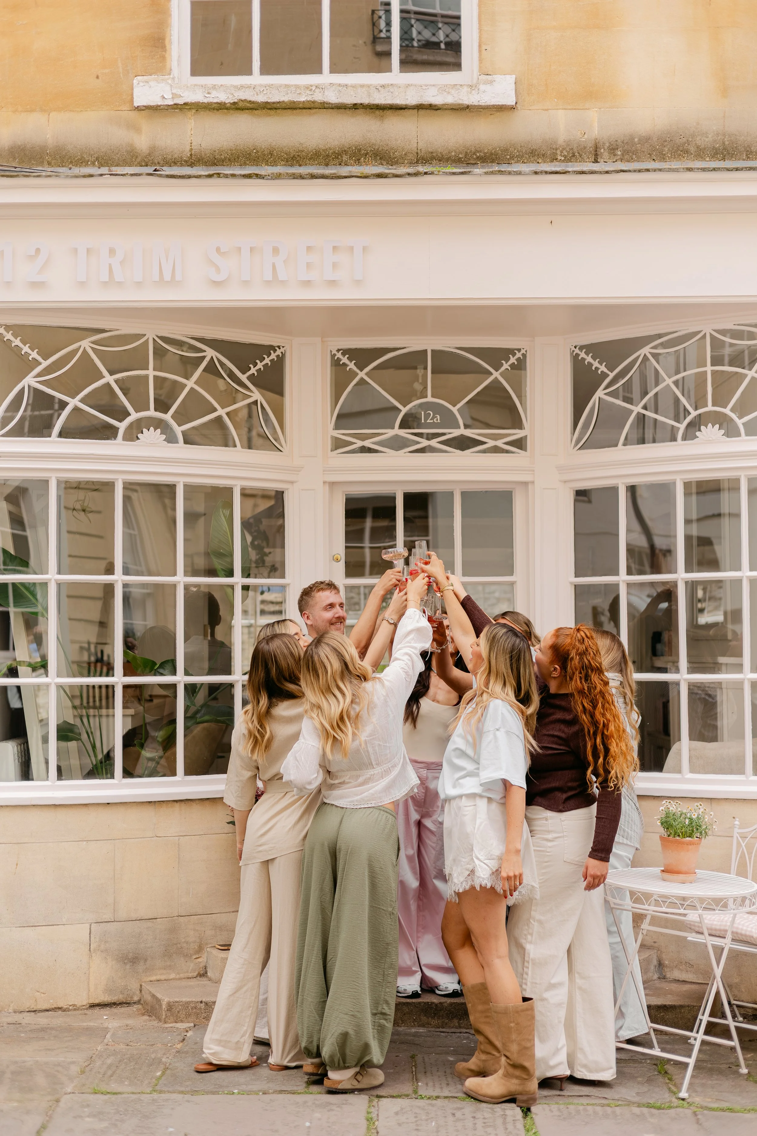 The team at 12 Trim Street raising glasses in a toast outside the salon in Bath with a sign reading '12 TRIM STREET' above a large window with decorative white frames.