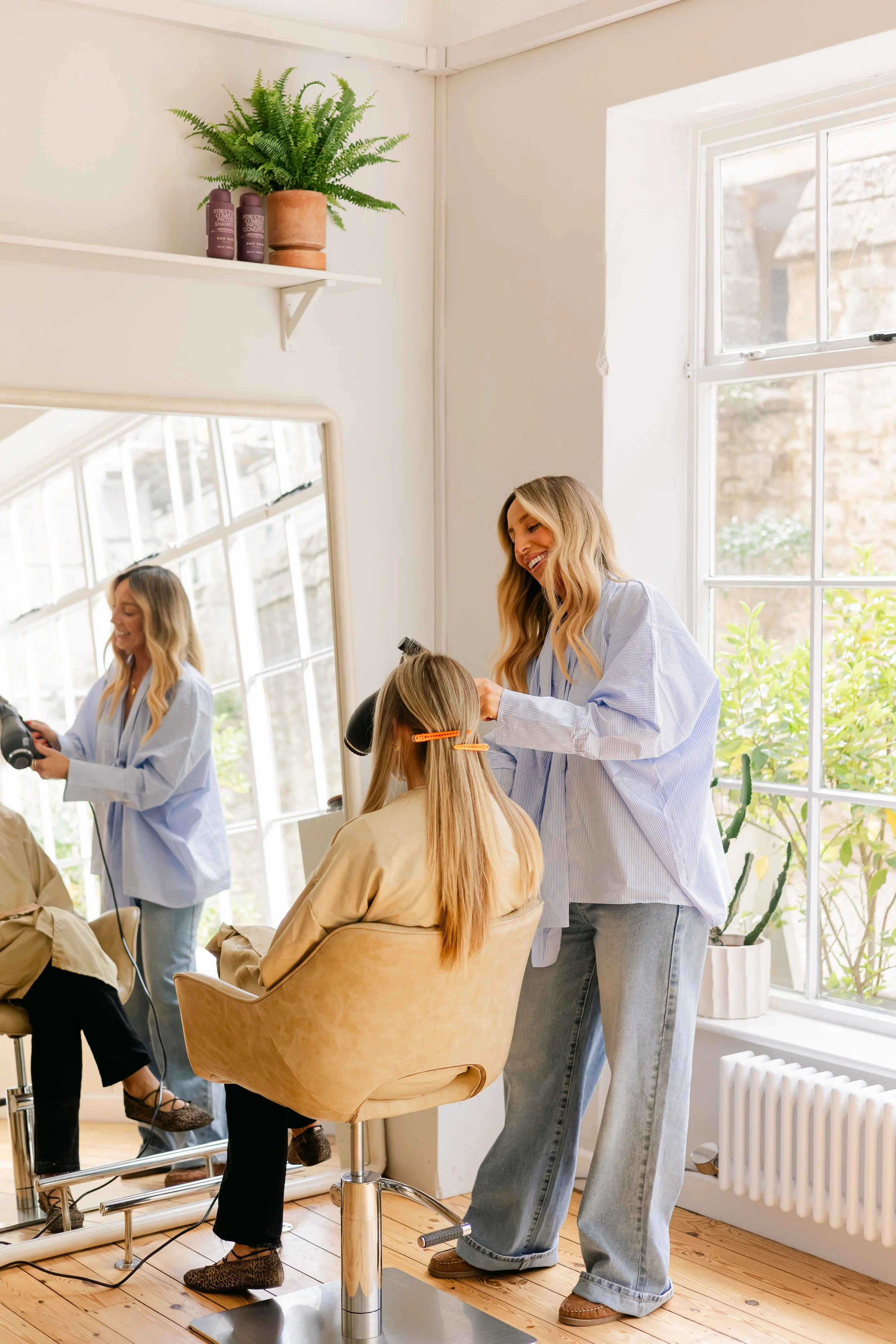 A hairstylist styling a woman’s hair in a bright, airy salon with large windows and a wooden floor.
