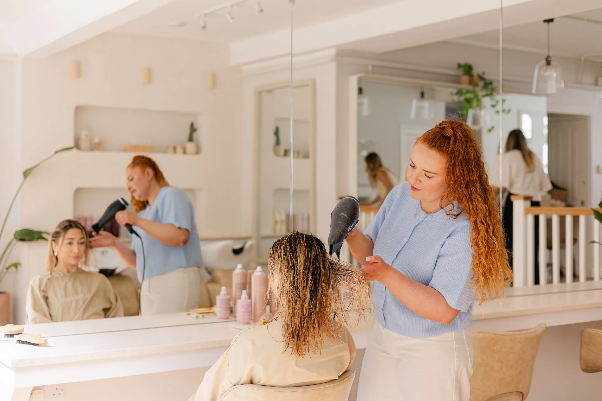 A hairstylist blow-drying a woman's hair in a salon with a mirror, other clients, and salon decor visible.