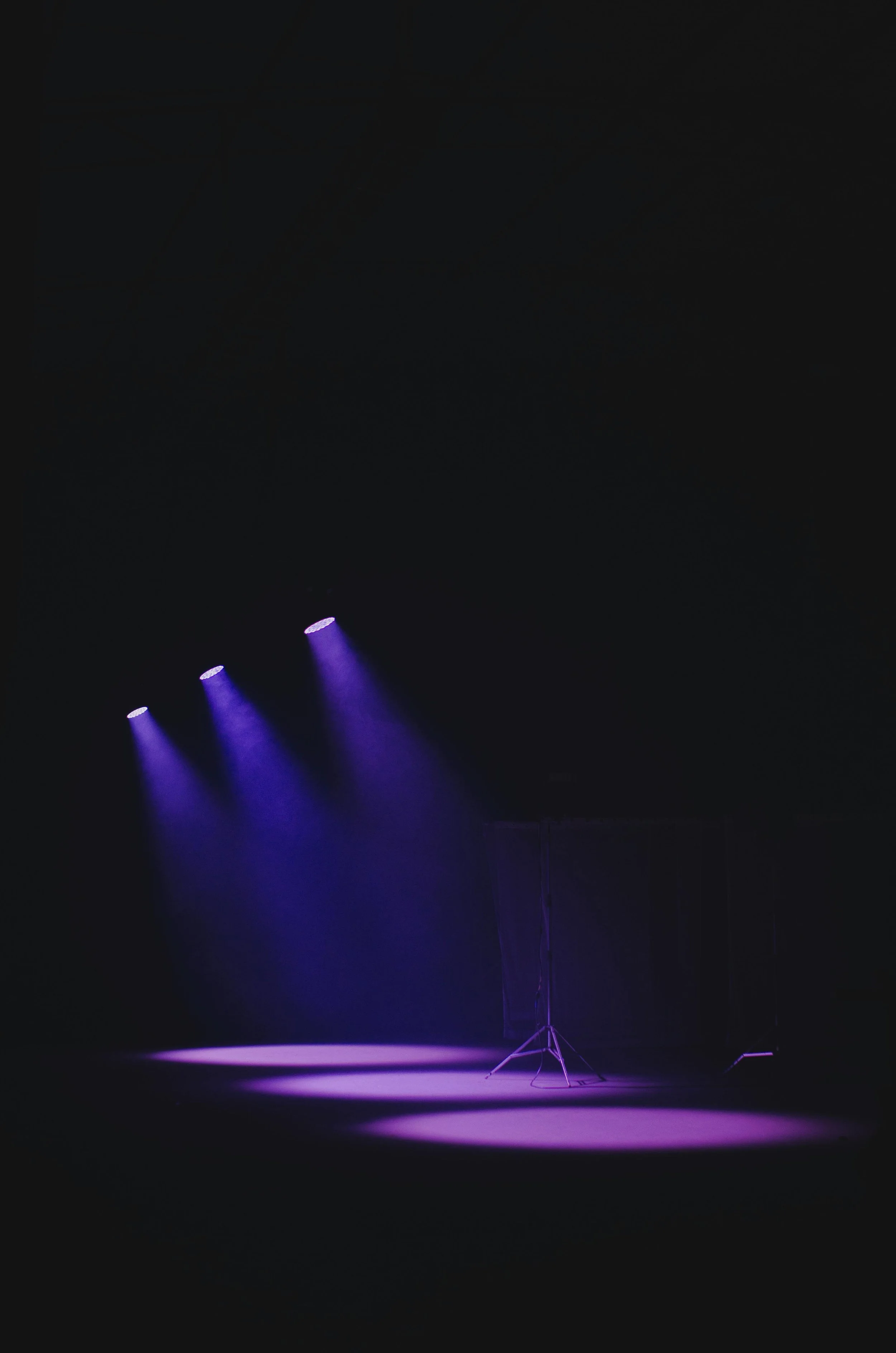 Empty stage with three purple spotlights shining down, spotlight stand and curtain in the background.