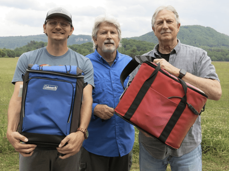 Three men standing outdoors in a grassy field with hills in the background, holding colorful cooler bags.