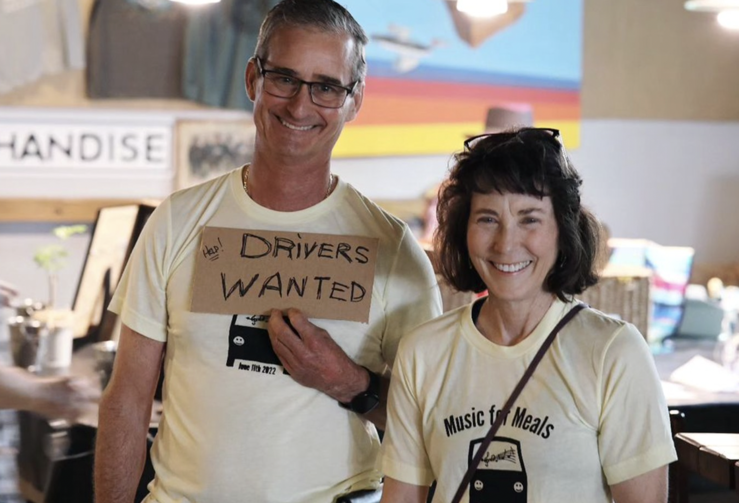 A man and a woman smiling indoors, holding a sign that says 'Drivers Wanted'. They are wearing yellow T-shirts with a logo that reads 'Music for Meals'.