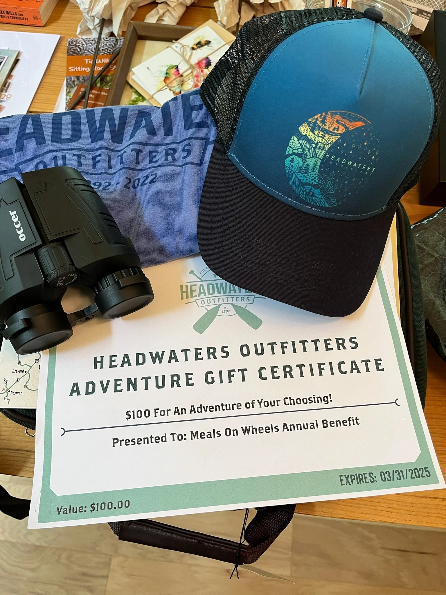 A gift certificate from Headwaters Outfitters for $100, with a cap and binoculars placed on top, on a table with various items in the background.