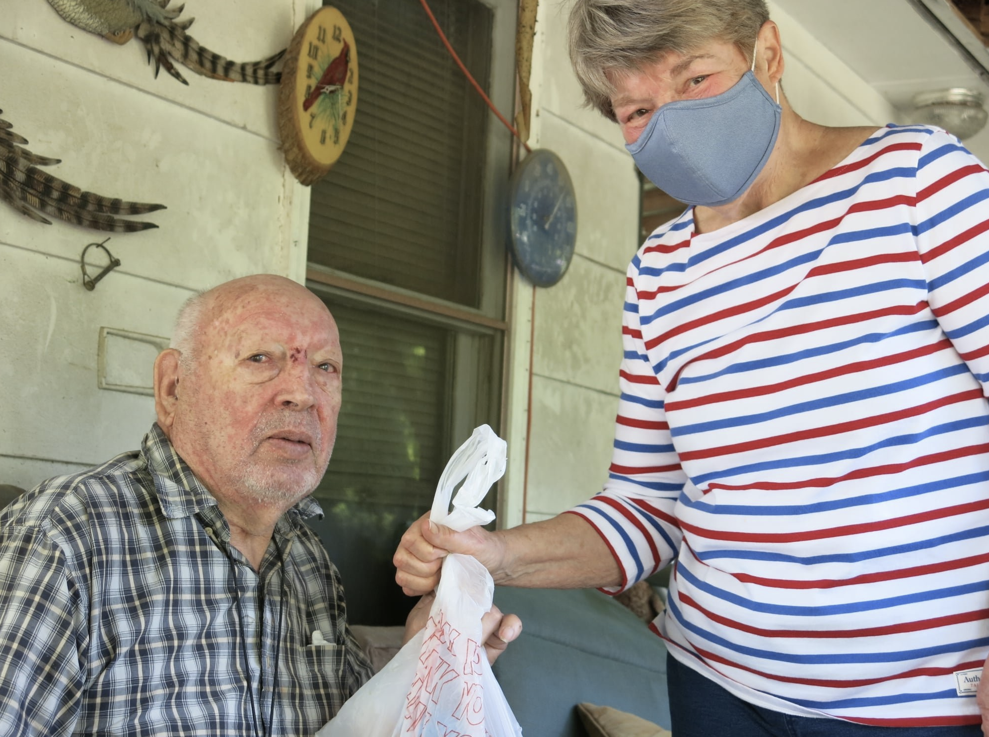 An elderly man sitting outdoors with a serious expression, wearing a plaid shirt. A woman with gray hair, wearing a face mask and a red, white, and blue striped shirt, is handing him a plastic bag, possibly containing groceries or food.