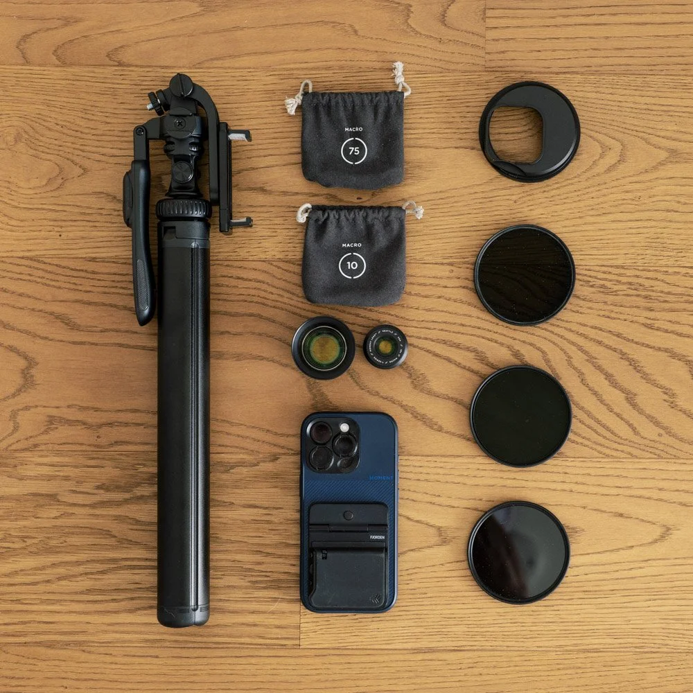 Fotografie von Fotografie-Equipment auf einem Holztisch, einschließlich eines Kamera-Stativs, vier Filterlinsen, zwei Aufbewahrungstaschen, einem Handy mit Kamera und einem Clip.