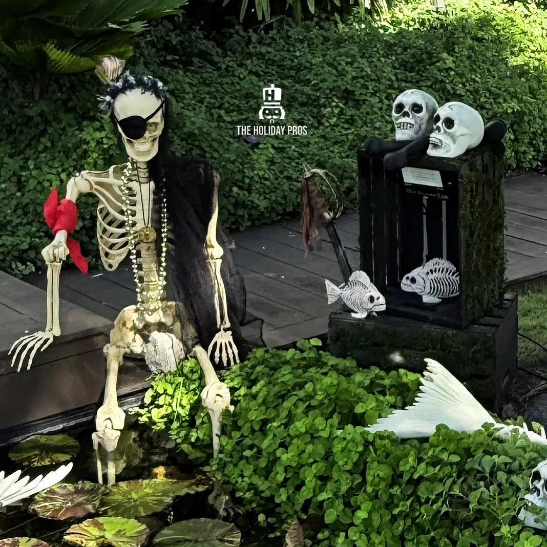 Skeleton dressed as a pirate with an eye patch and red bandana, sitting outdoors beside a pond with aquatic plants. Surrounding decorations include skulls on a black crate, fish skeletons, and a boat-shaped object resembling a fish or whale. The scene appears to be a Halloween or Day of the Dead display.