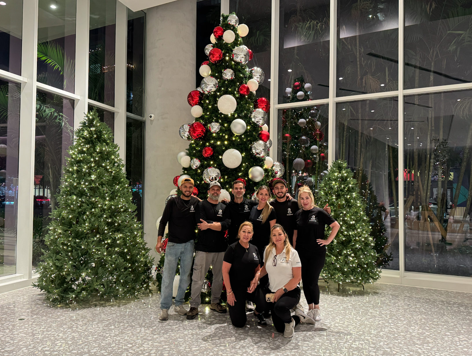 A group of nine people posing in front of a large decorated Christmas tree with three smaller trees, inside a building with floor-to-ceiling glass windows at night.