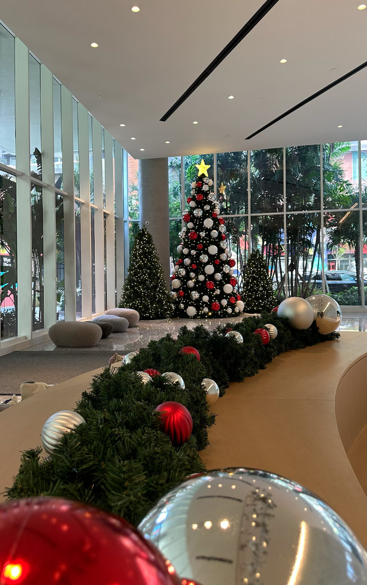 Decorated Christmas tree with red, white, and gold ornaments inside a modern building with large glass windows and Christmas garland.