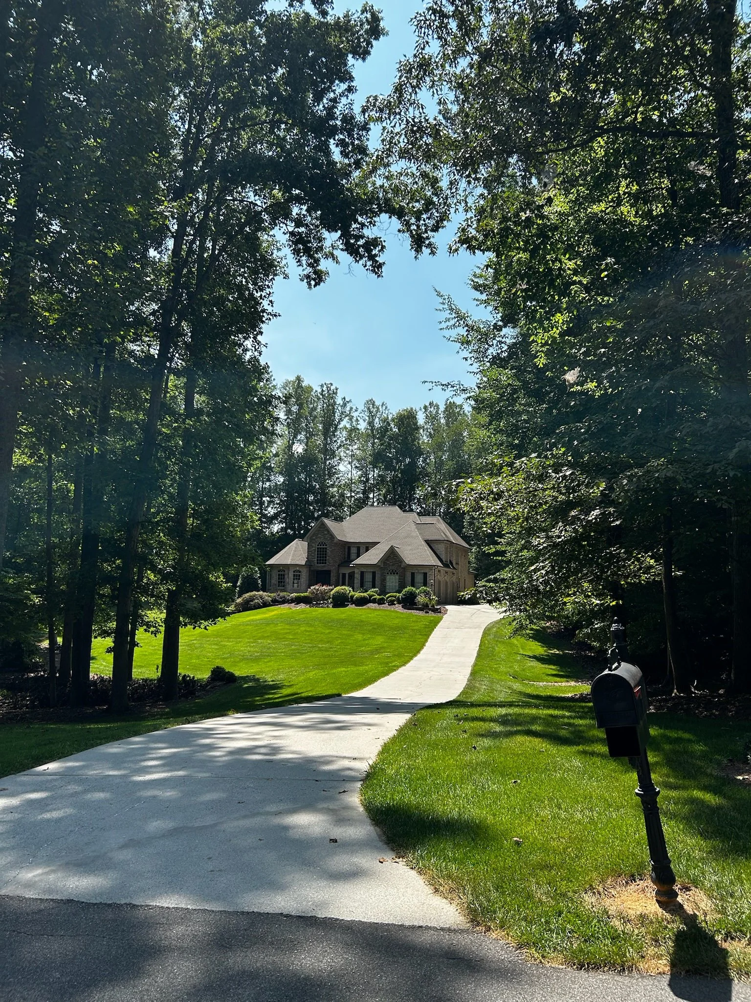 A large house at the top of a hill, with a curved driveway leading up to it, surrounded by green grass and trees, with a mailbox near the driveway.