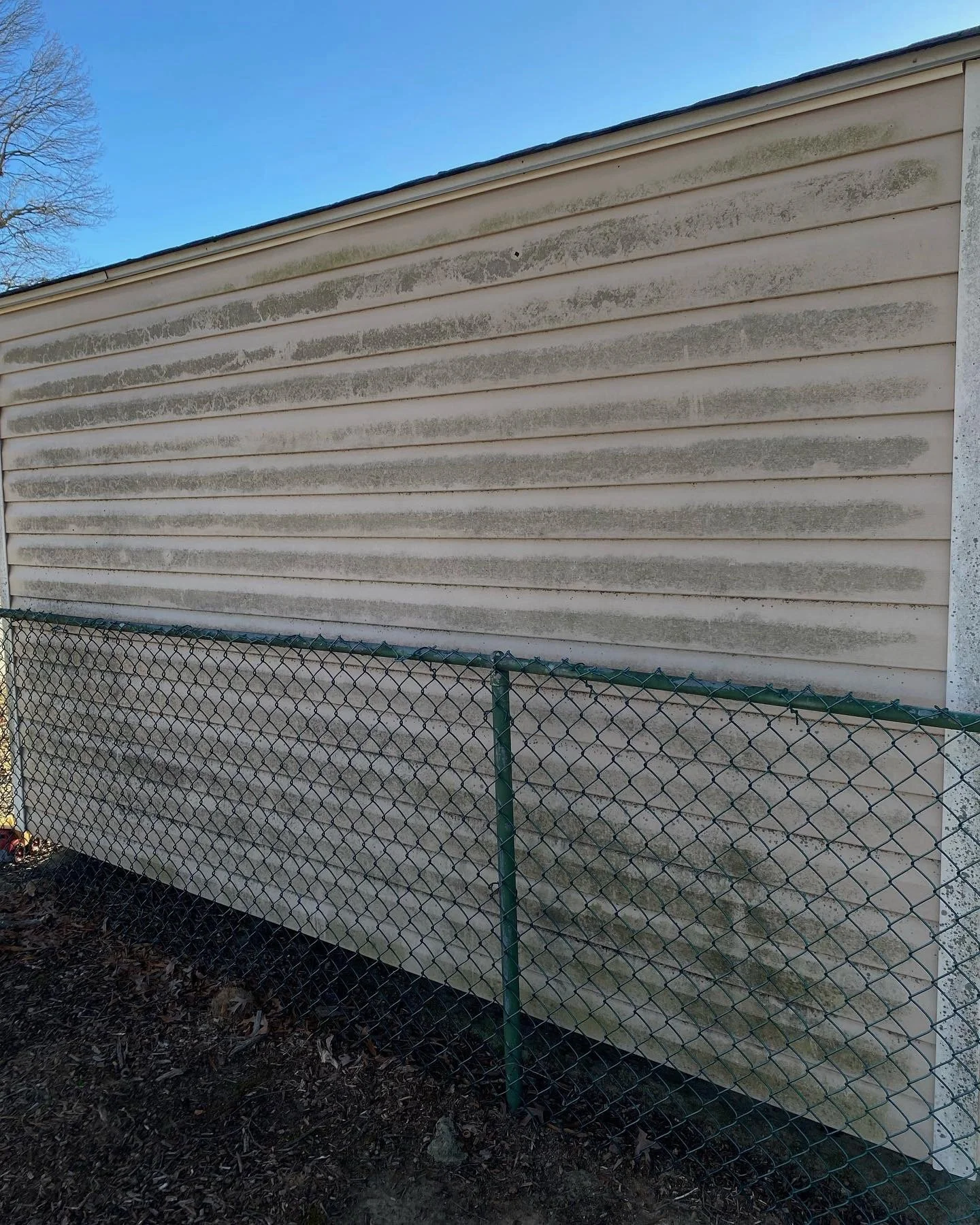 The exterior of a house with beige siding showing dirt, mold, or mildew streaks, and a chain-link fence in front.