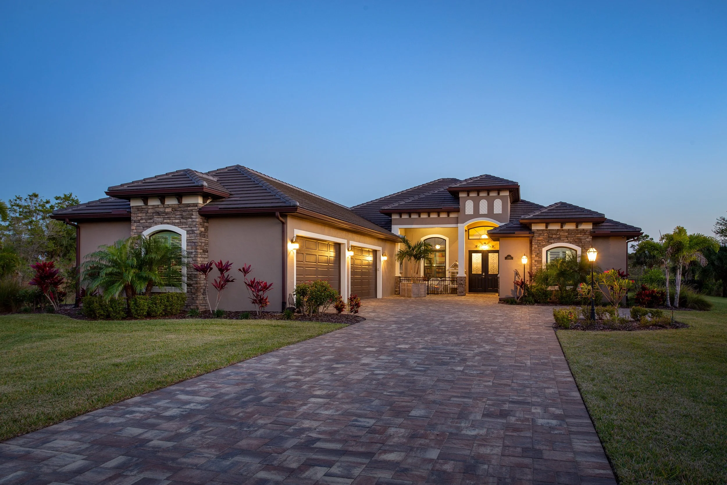 A Florida home with lights glowing at dusk with paver driveway and tropical landscaping