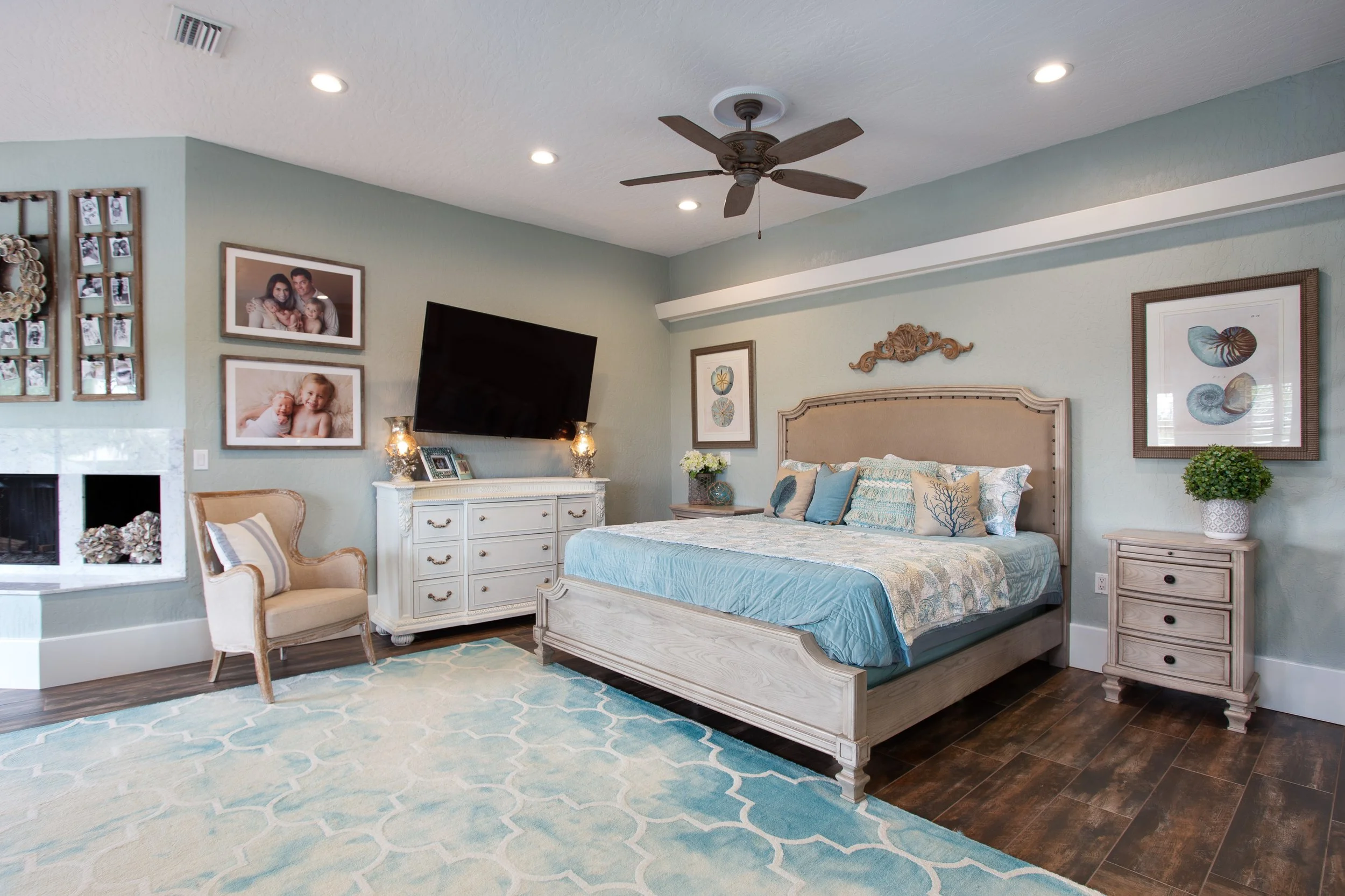 A cozy bedroom with a large bed, beige headboard, and matching nightstands. Decor includes framed artwork of seashells and coral, a bouquet of white flowers, and a potted green plant. There's a white dresser with a wall-mounted flat-screen TV, a beige armchair, and family photos on the wall. The room has wood flooring, a light blue and white patterned rug, and overhead lighting with a ceiling fan.