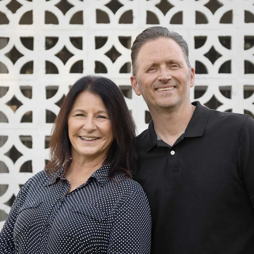 A smiling woman and man standing outdoors in front of a decorative white wall pattern.