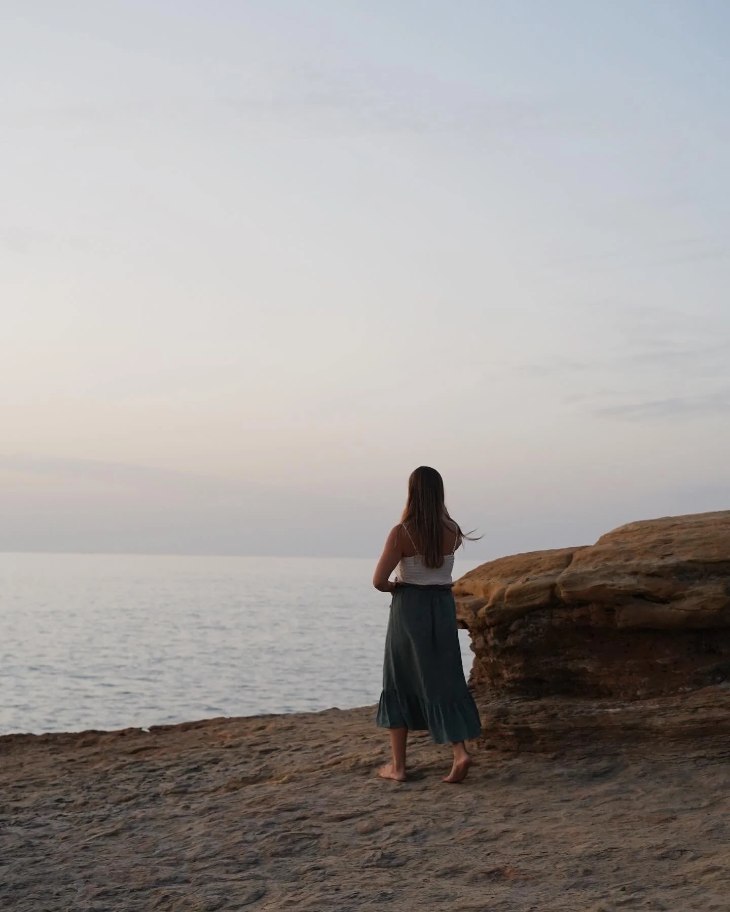 Woman standing on rocky shore overlooking calm sea during sunset.