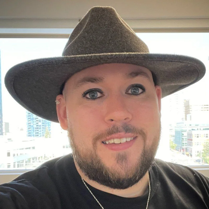 A man with a beard and makeup wearing a large cowboy hat and a black t-shirt, taking a selfie indoors with a cityscape visible through a window in the background.