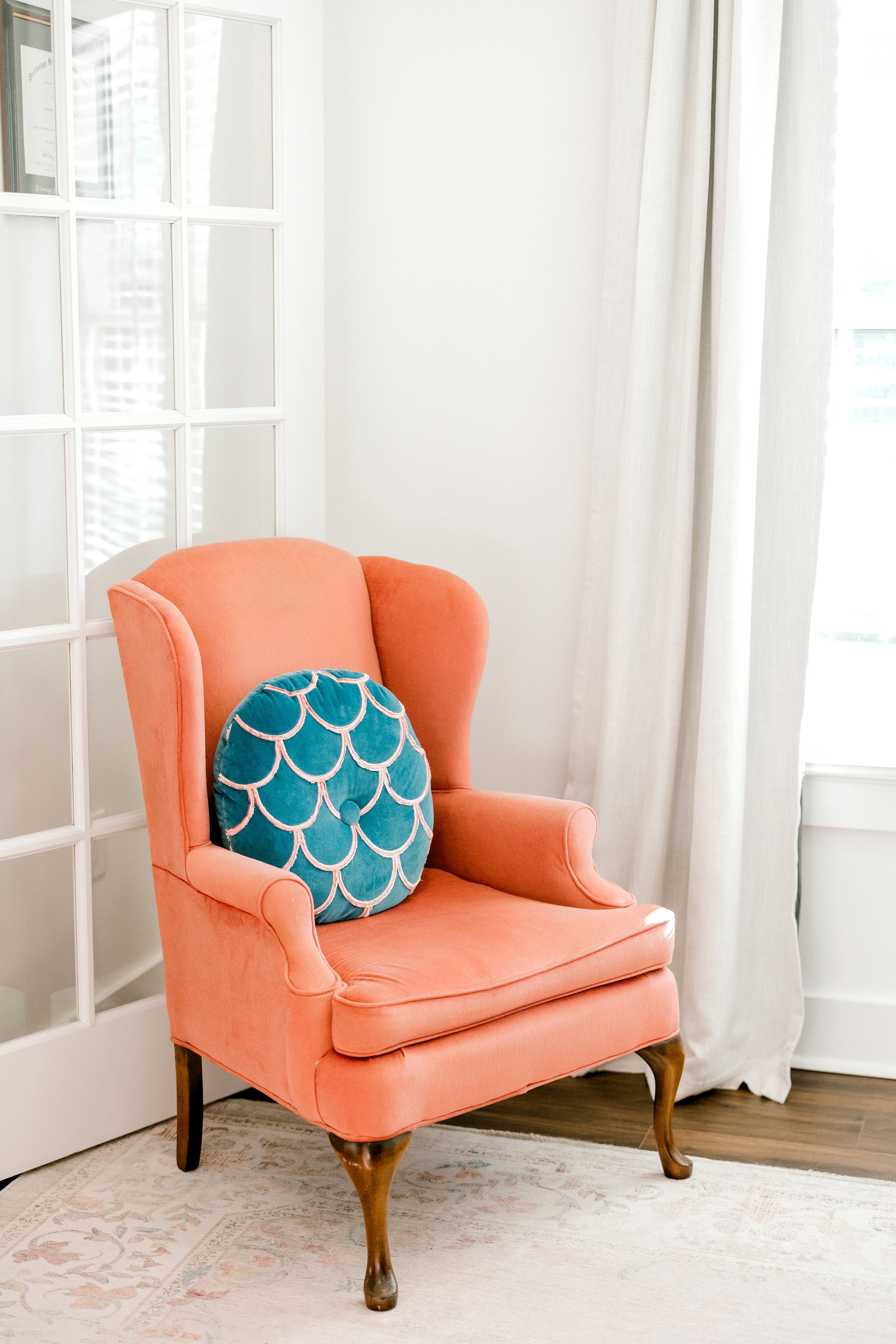 A pink upholstered armchair with wooden legs, decorated with a round teal pillow with pink scalloped embroidery, in a bright room with white walls, a white curtains, and a glass-paneled white door.