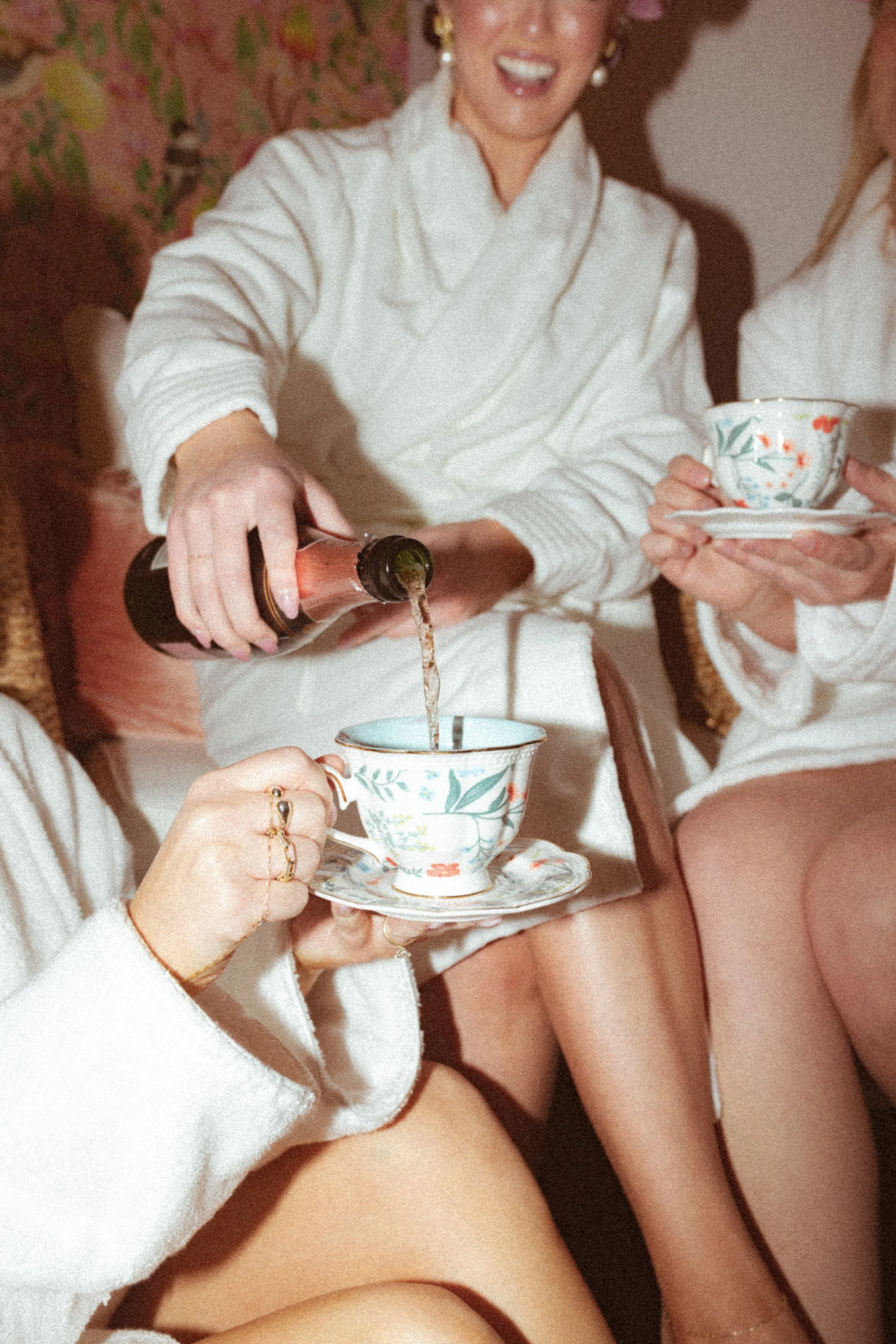 Two women in robes and towels on their heads smiling as one pours champagne into a glass in a bedroom.