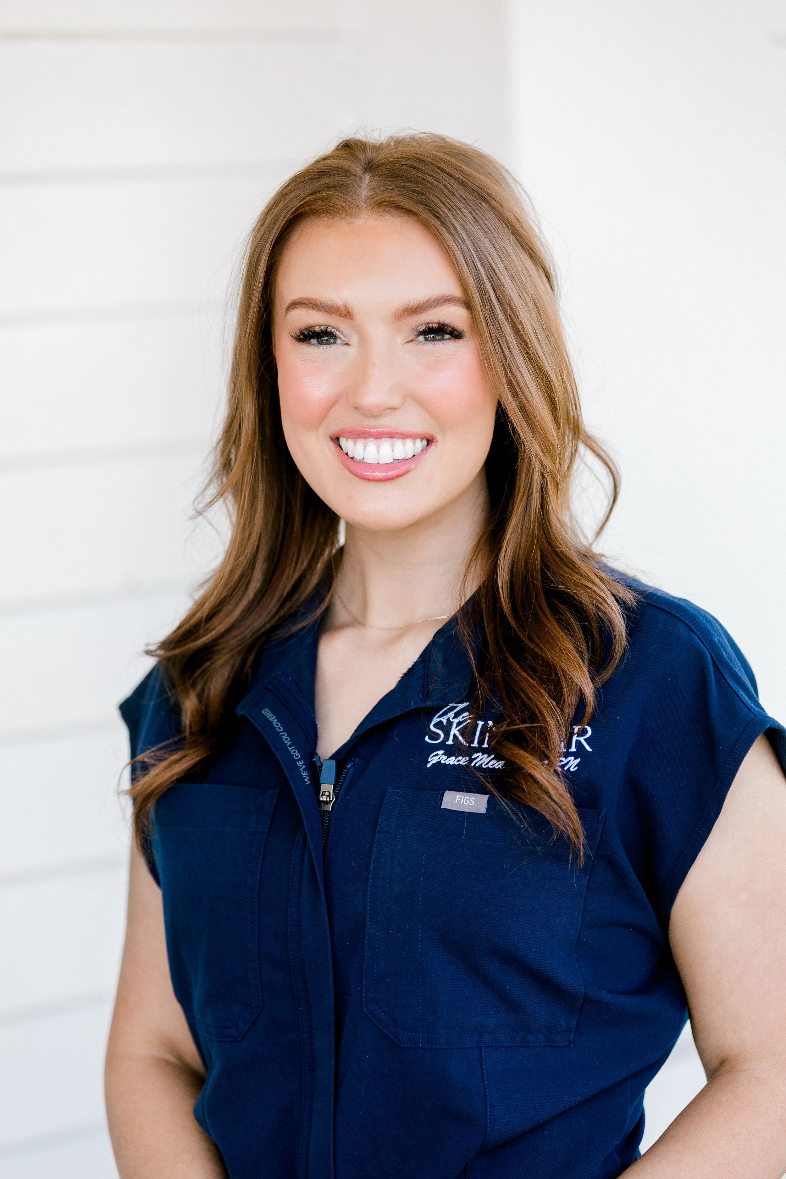 A woman with long, wavy reddish-brown hair, smiling, wearing a light blue shirt, outdoors with a blurred background of greenery and a white arch.