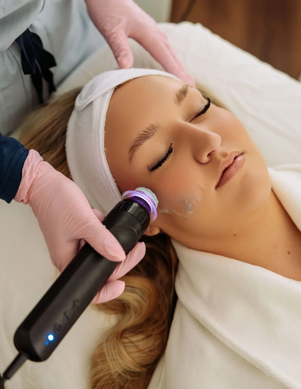A woman with long hair and a headband receiving a facial treatment using a device with a purple light, while lying on a treatment bed with her eyes closed.