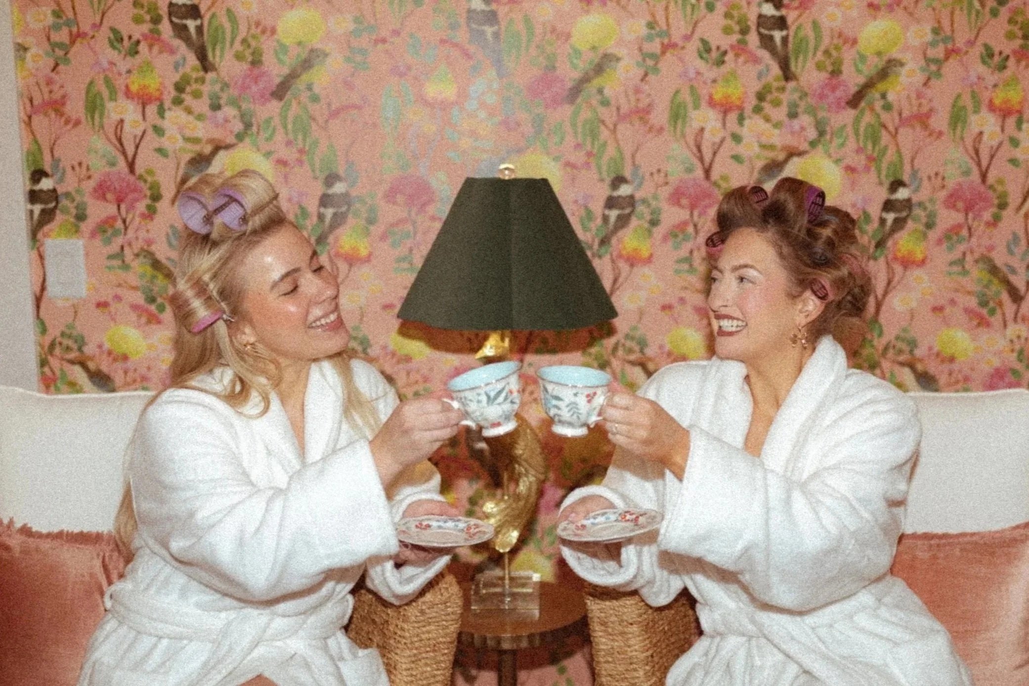 Two women in white robes and towels on their heads sitting on a bed, holding glasses of champagne, smiling and laughing together.