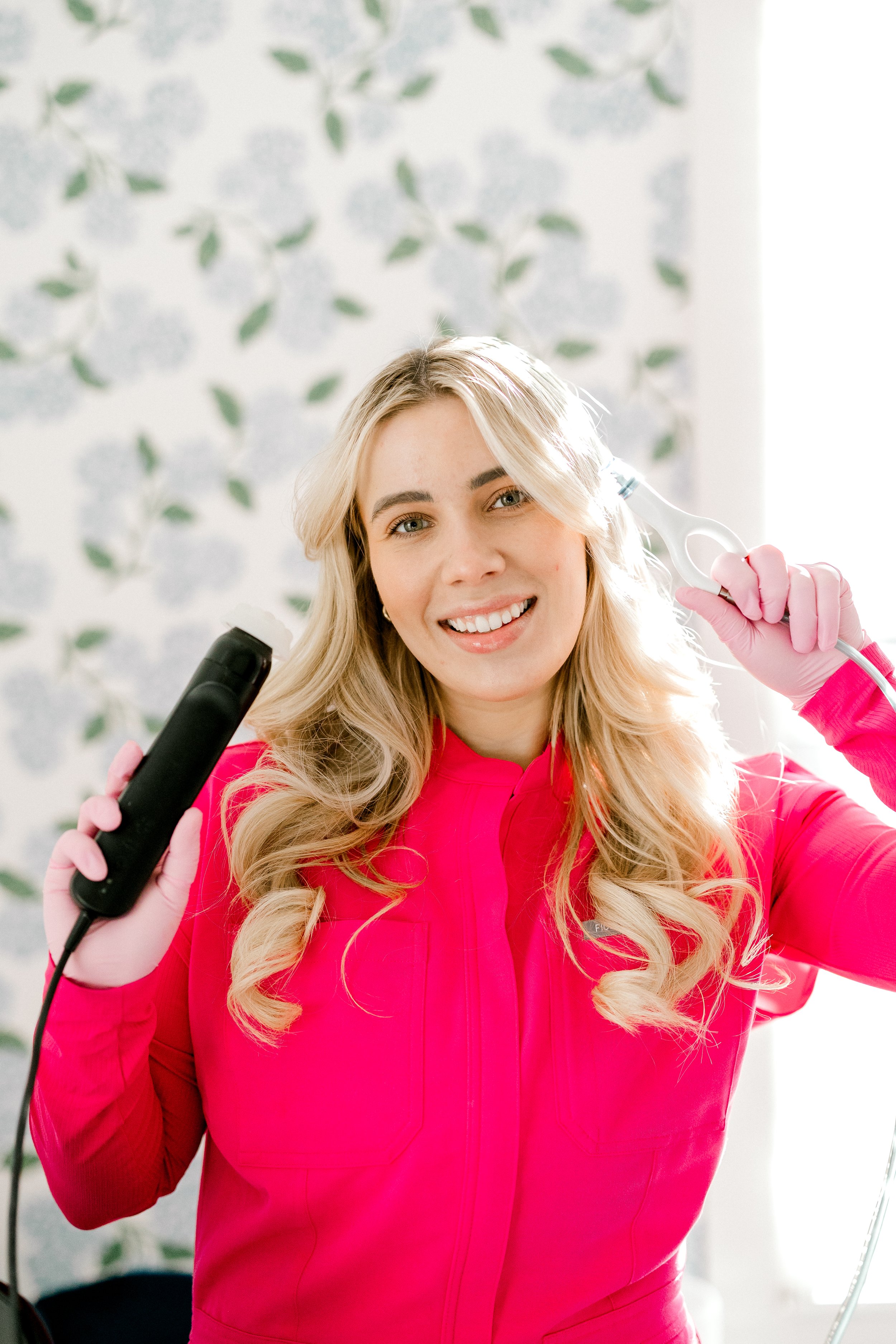 A woman with blonde hair smiling, holding a curling iron in one hand and a flat iron in the other, wearing a bright pink top and pink gloves, standing in front of a floral-patterned wall.