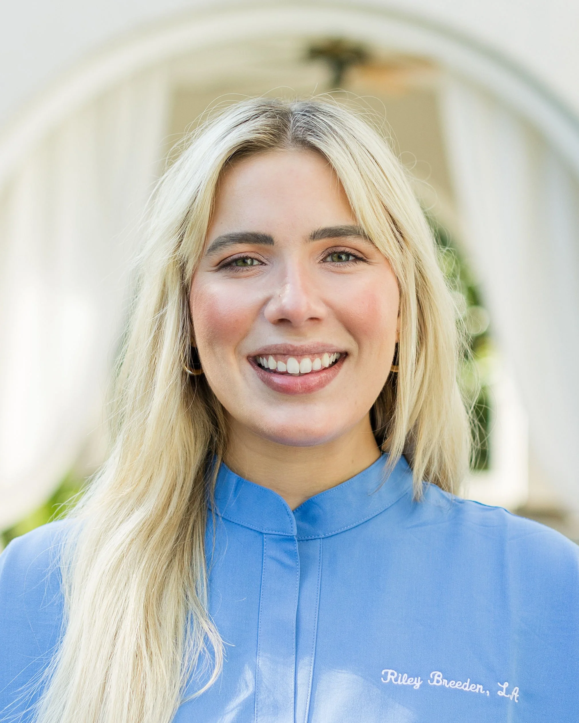 Close-up of a woman with long blonde hair smiling, wearing a light blue veterinarian coat with embroidered name 'Riley Breeden, LV' standing in a bright, clean indoor space.