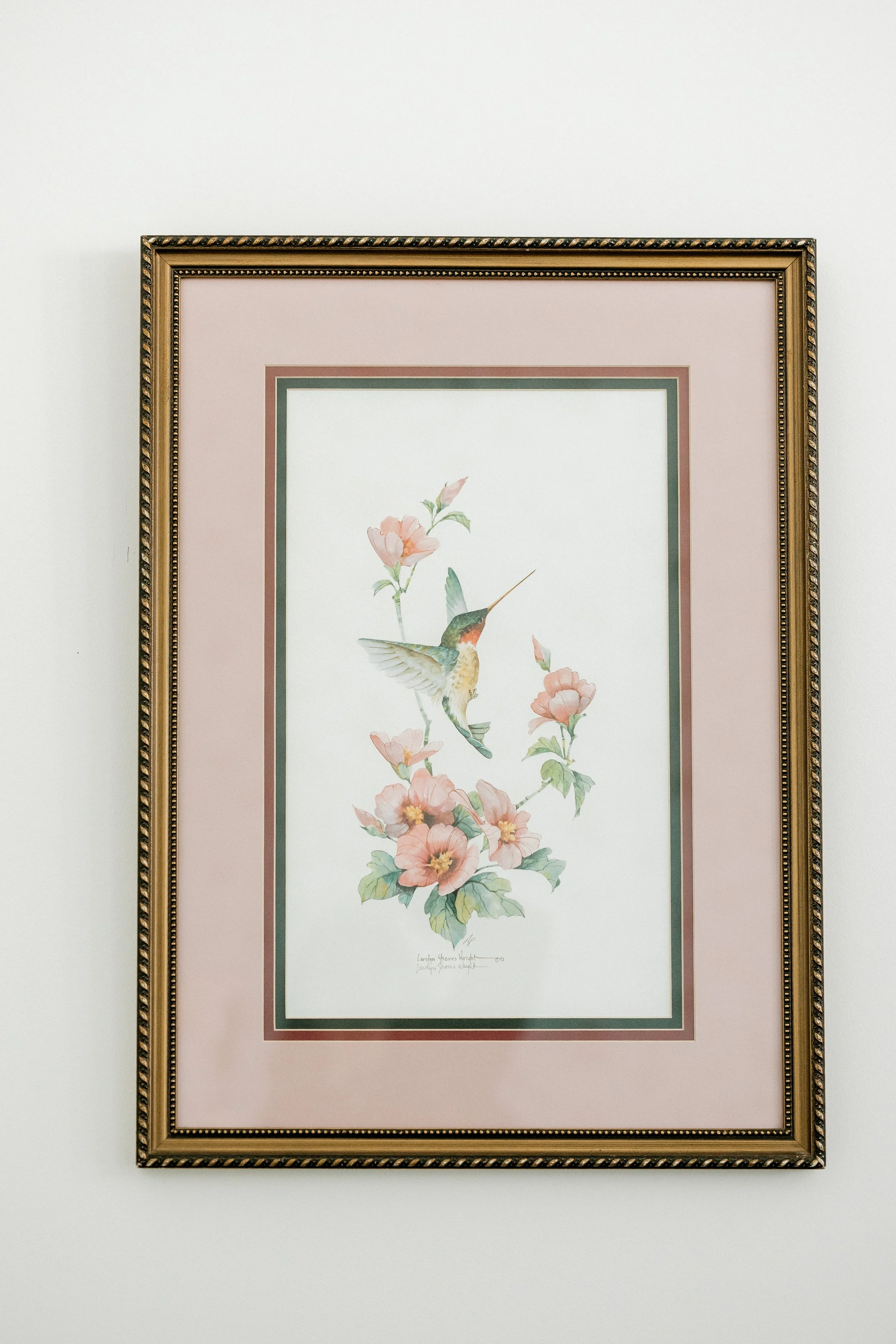 Framed watercolor painting of a hummingbird flying among pink flowers on a white wall.