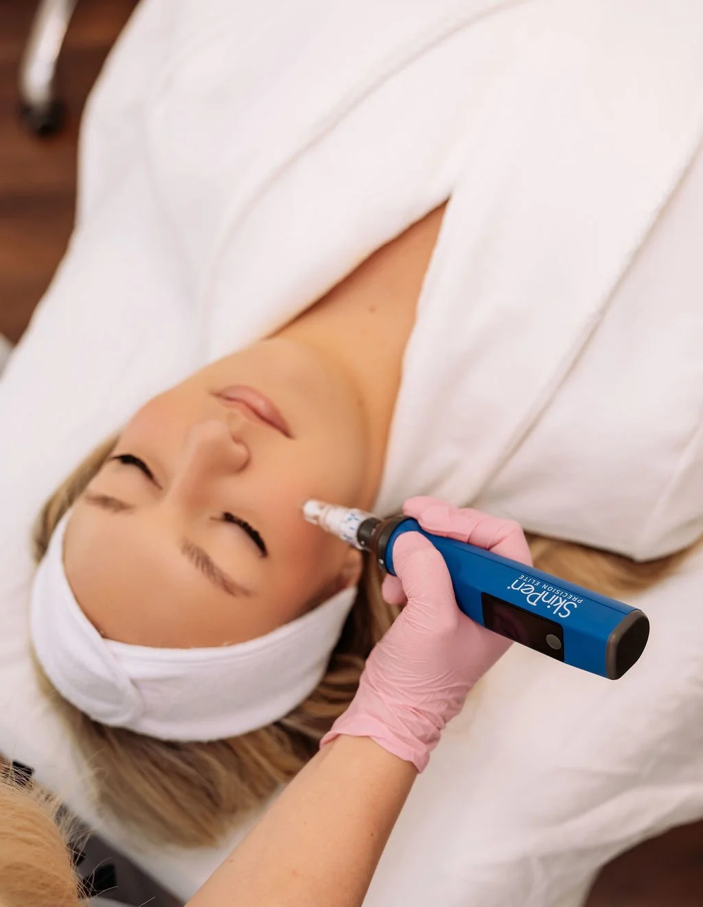 A woman lying down with eyes closed as a technician administers a facial treatment with a SkinPen microneedling device on her cheek. The woman is wearing a white headband and a white robe.