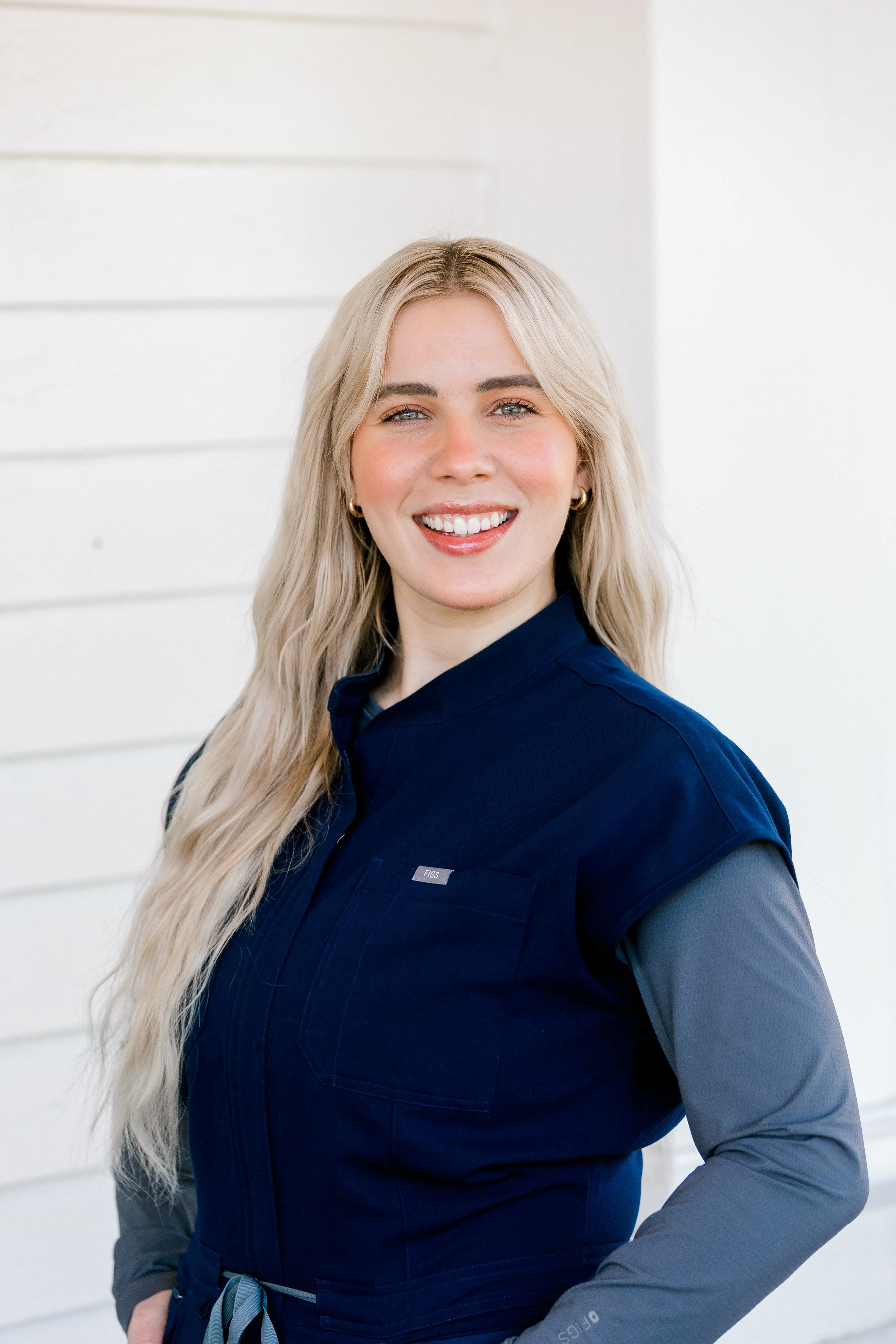 Close-up of a woman with long blonde hair smiling, wearing a light blue veterinarian coat with embroidered name 'Riley Breeden, LV' standing in a bright, clean indoor space.