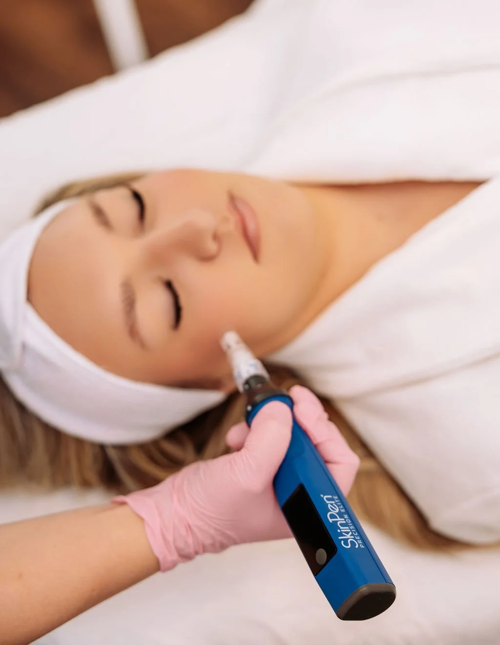 A woman receiving a cosmetic treatment with a SkinPen microneedling device on her cheek, lying on a bed with a white headband and white sheets.
