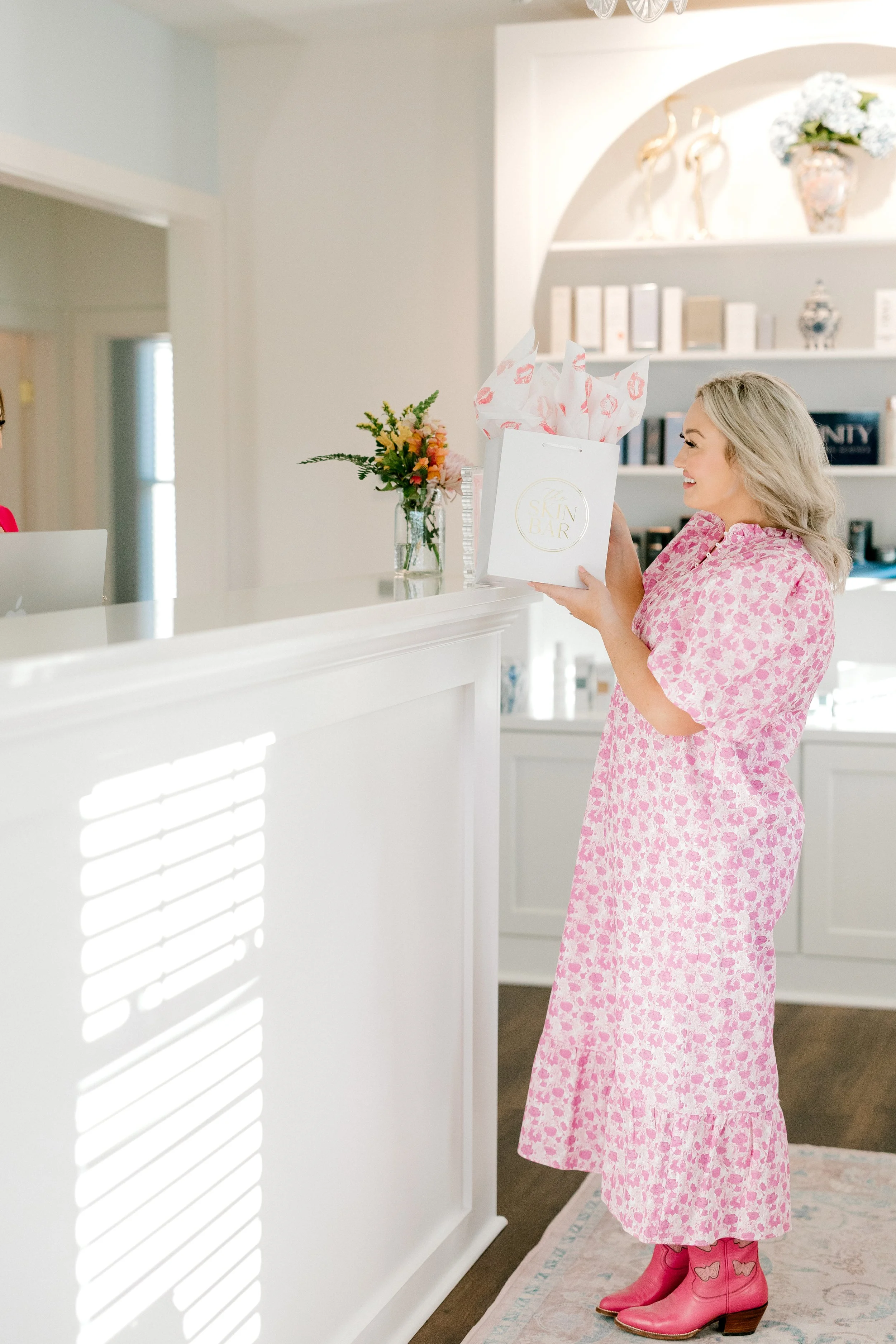 A woman receiving a gift while standing at a reception counter in a bright, white-themed room. She is wearing a pink patterned dress and pink cowboy boots, smiling as she holds a white shopping bag with tissue paper.