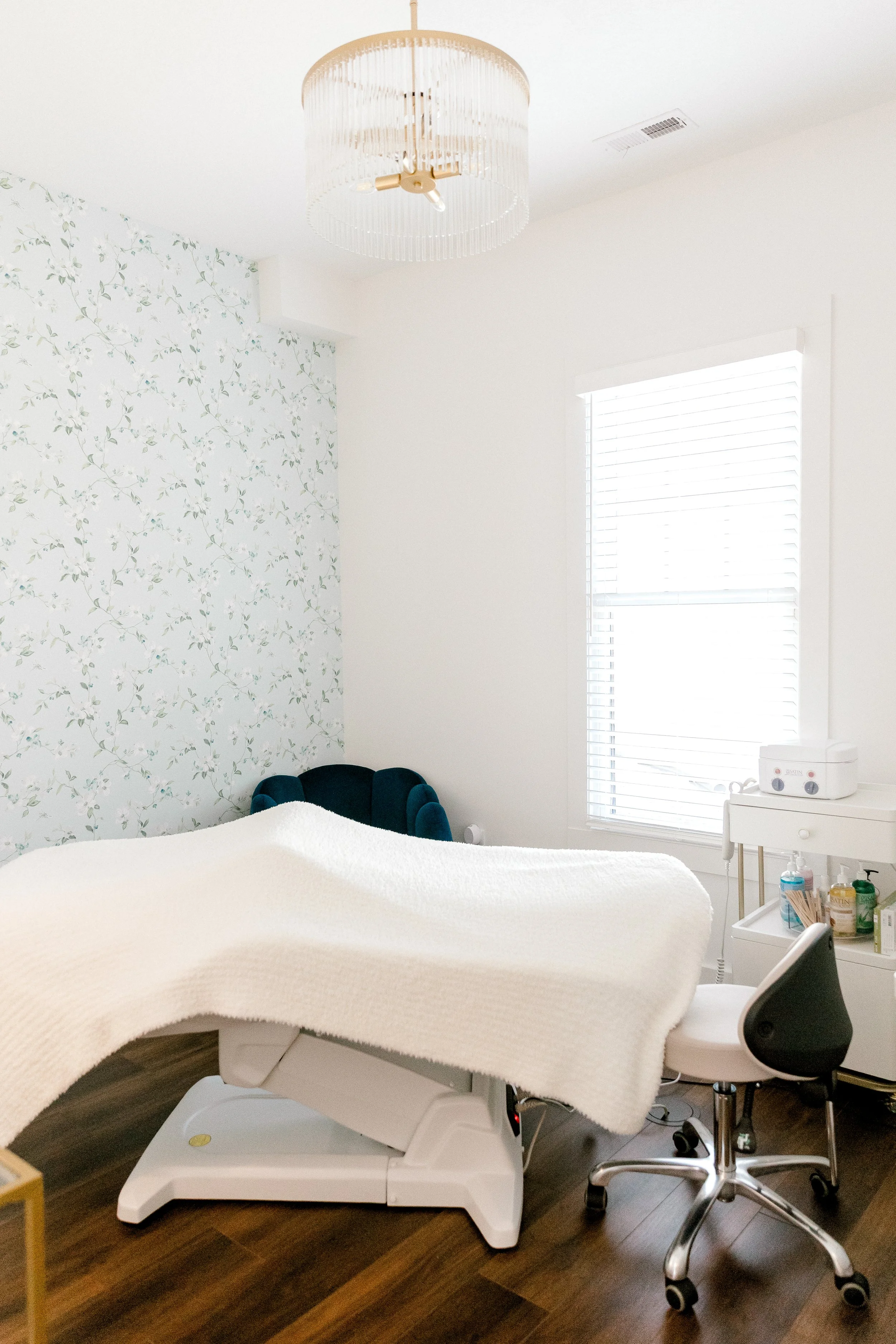 Medical examination room with a patient bed, a blue armchair, a rolling chair, and a white cabinet with medical supplies, illuminated by a chandelier and a window with blinds.