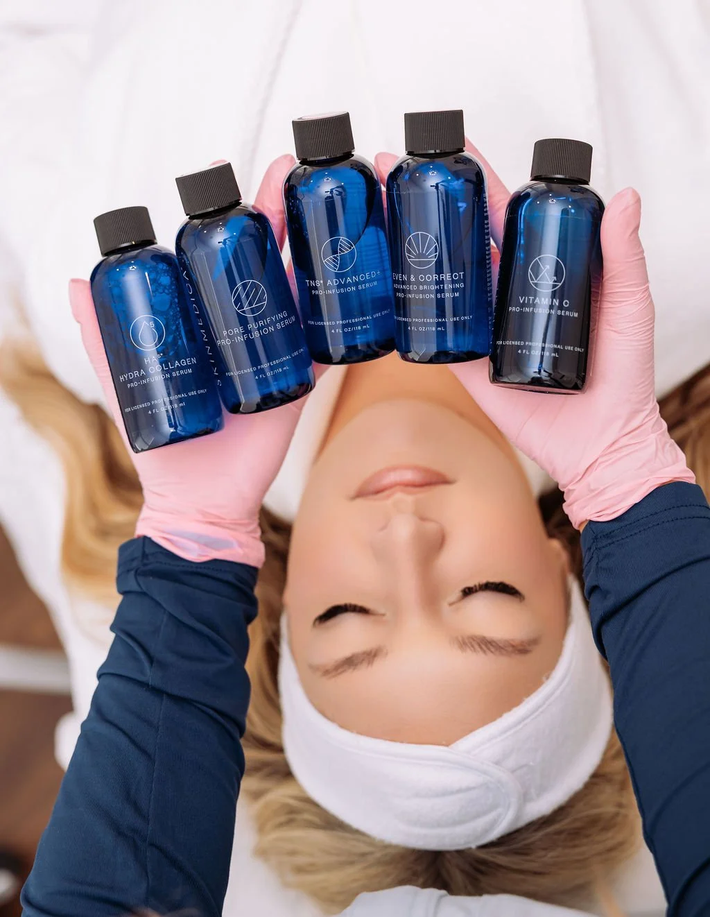 A woman wearing pink gloves and a white headband lying down with her eyes closed, holding five blue bottles of skincare serums labeled 'Hydra Collagen', 'Pore Purifying', 'Advanced Brightening', 'Even & Correct', and 'Vitamin C' in a beauty or skincare setting.