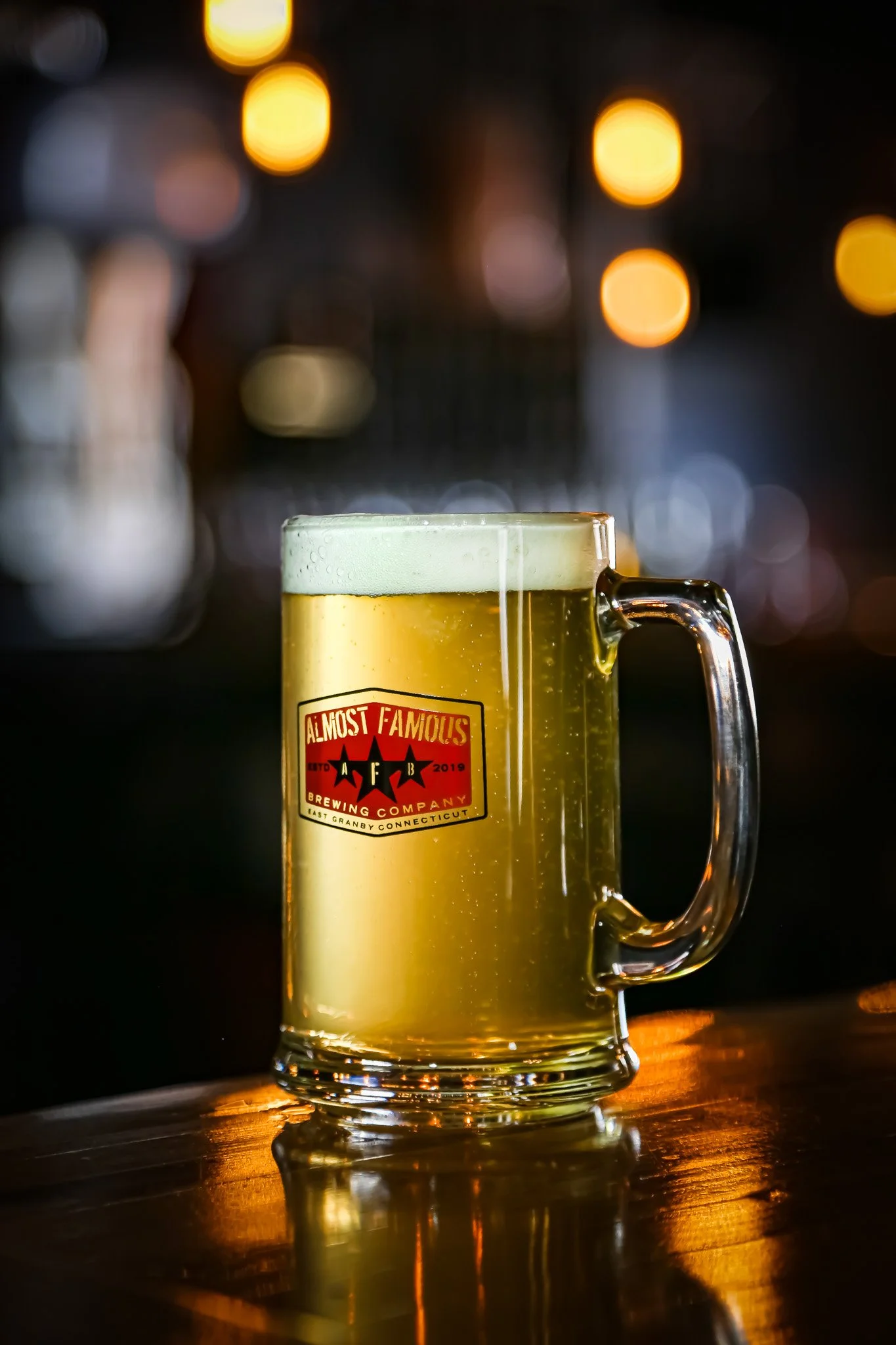Glass mug of beer with "Almost Famous Brewing Company" label, on a wooden bar surface with bokeh lights in the background.