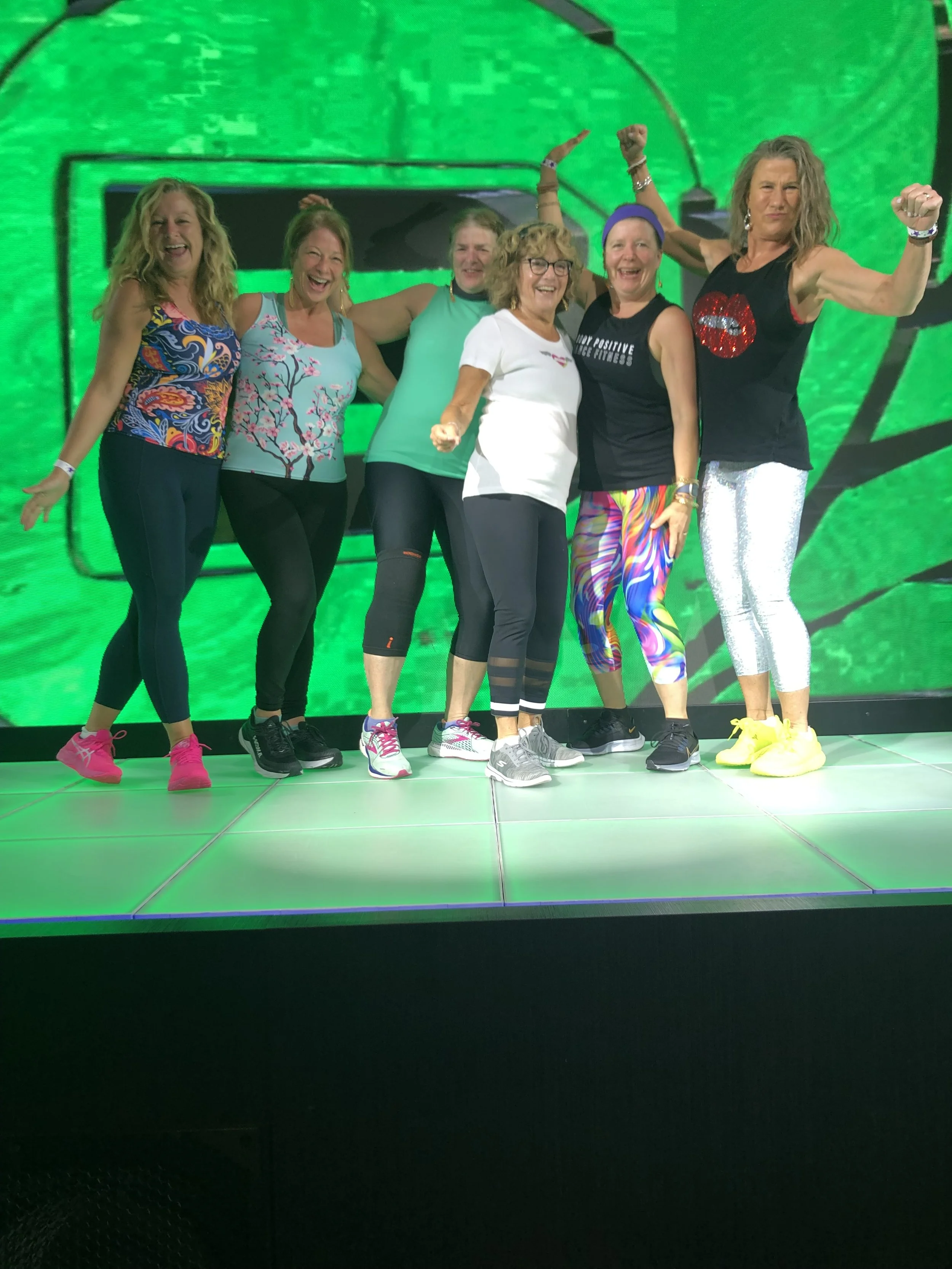 A group of seven women on stage in activewear, smiling and celebrating with a green digital background.