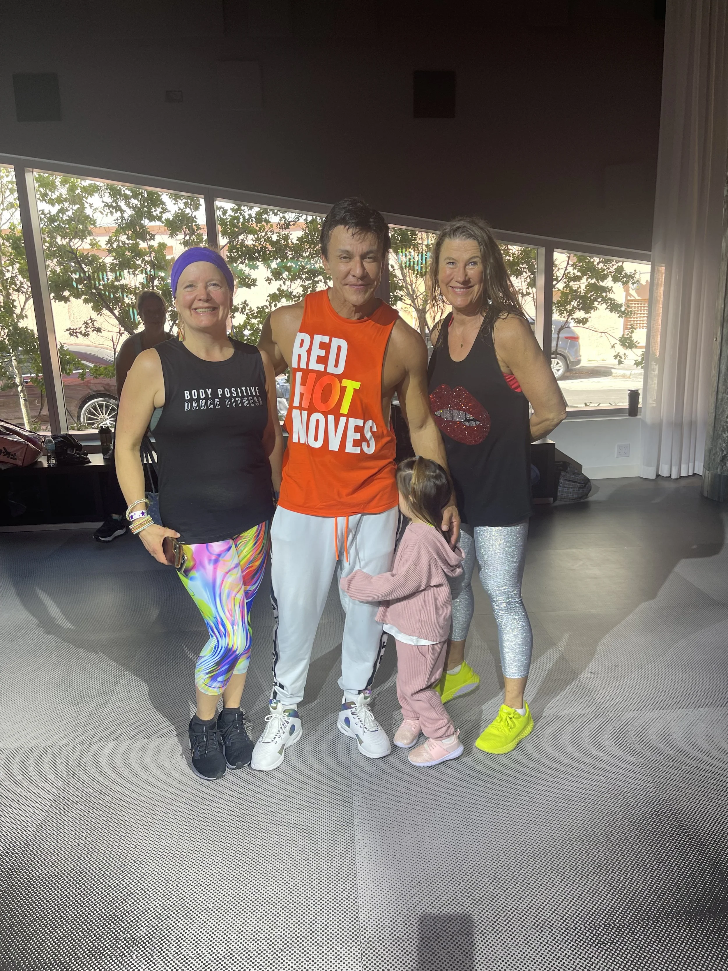 Group of four people, three adults and one child, standing together indoors, wearing athletic clothing, smiling at the camera.