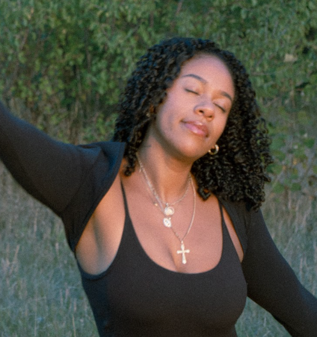 A woman stands outdoors with eyes closed, raising her arm in a peaceful gesture, surrounded by greenery.