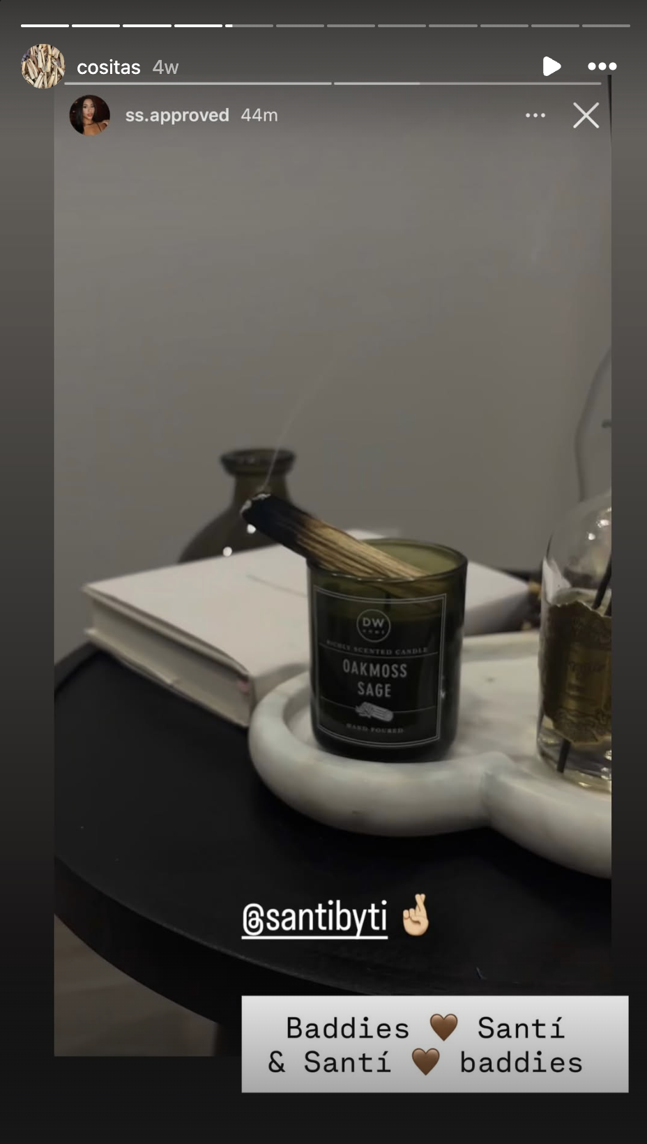 A white marble tray holding a black Oakmoss Sage scented candle, a clear glass bottle with a herbal infusion, a closed notebook, and a white object, all on a black surface with a gray wall background.