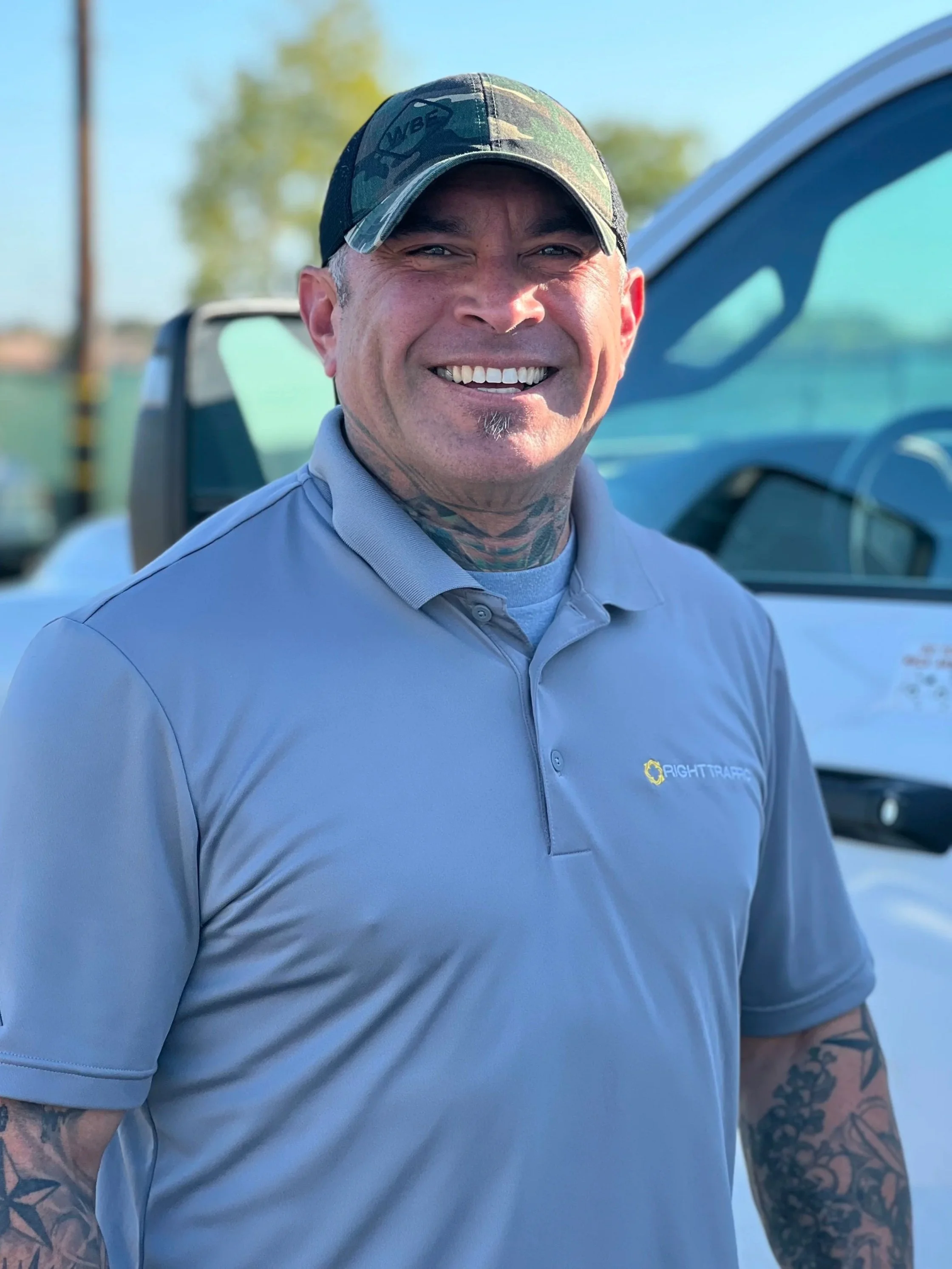A man smiling outdoors, wearing a gray collared shirt with 'Right Traff' logo, a camouflage baseball cap, and showing tattoos on his neck and arms, with a blue vehicle in the background.