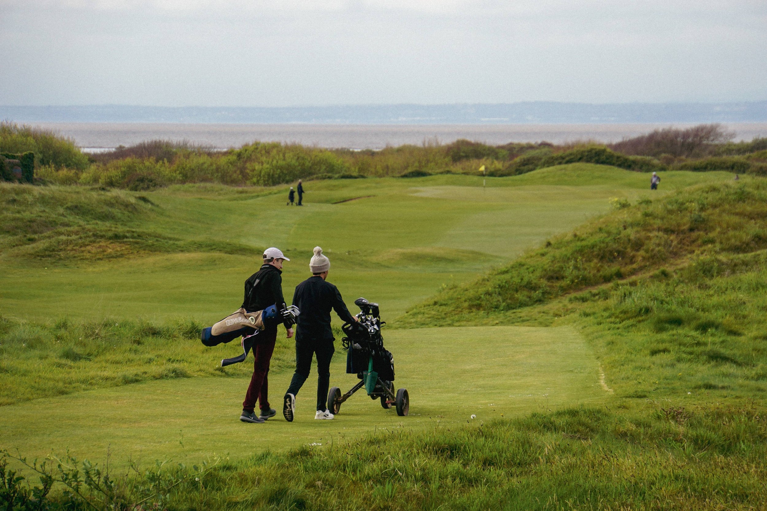 Two people walking on a golf course carrying golf bags, with a view of the green and flag, surrounded by grassy hills and a body of water in the distance.