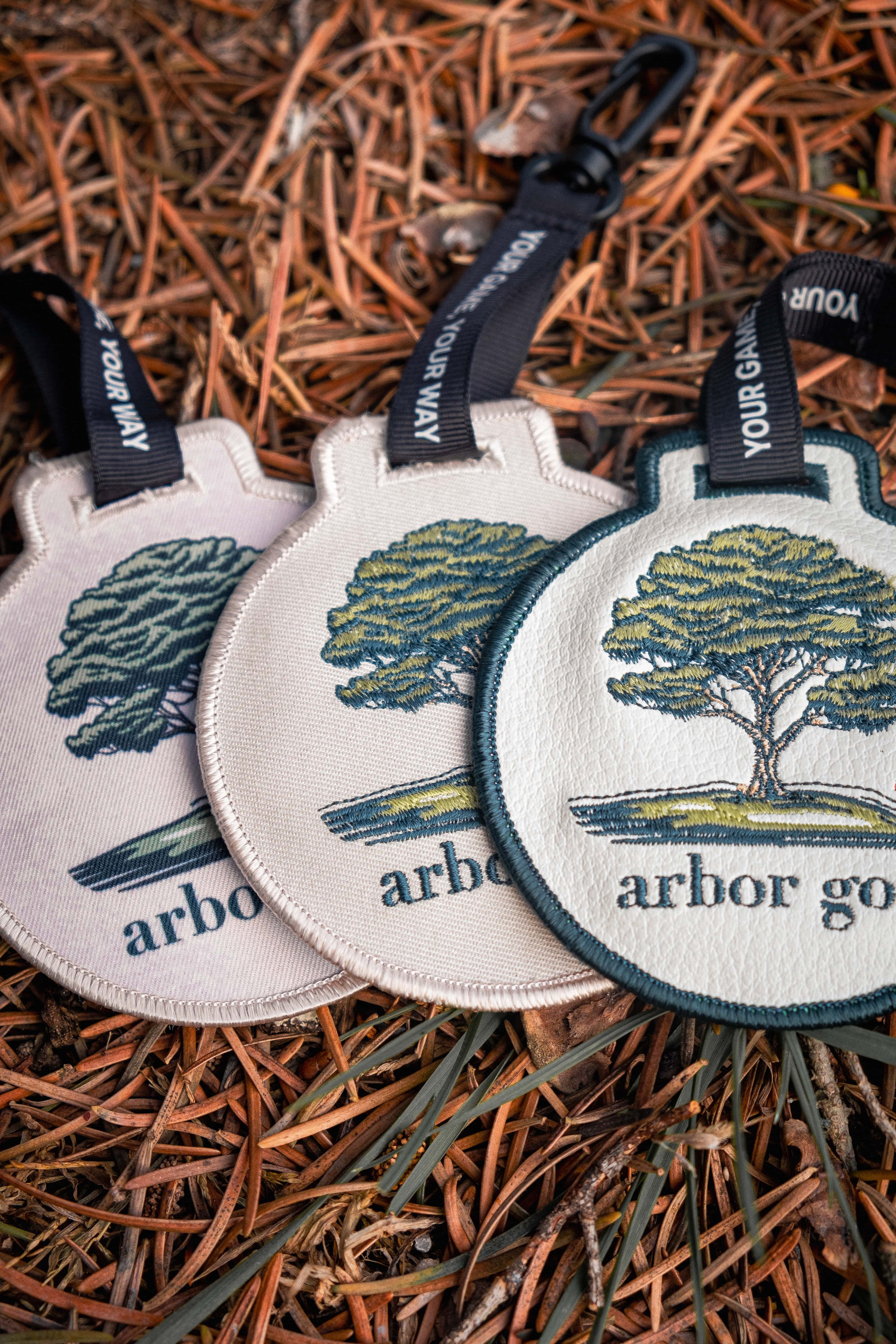 Close-up of three round, embroidered golf bag tags with tree logo, labeled "arbor golf," placed on a bed of pine needles. Each tag has a black strap with a clip.