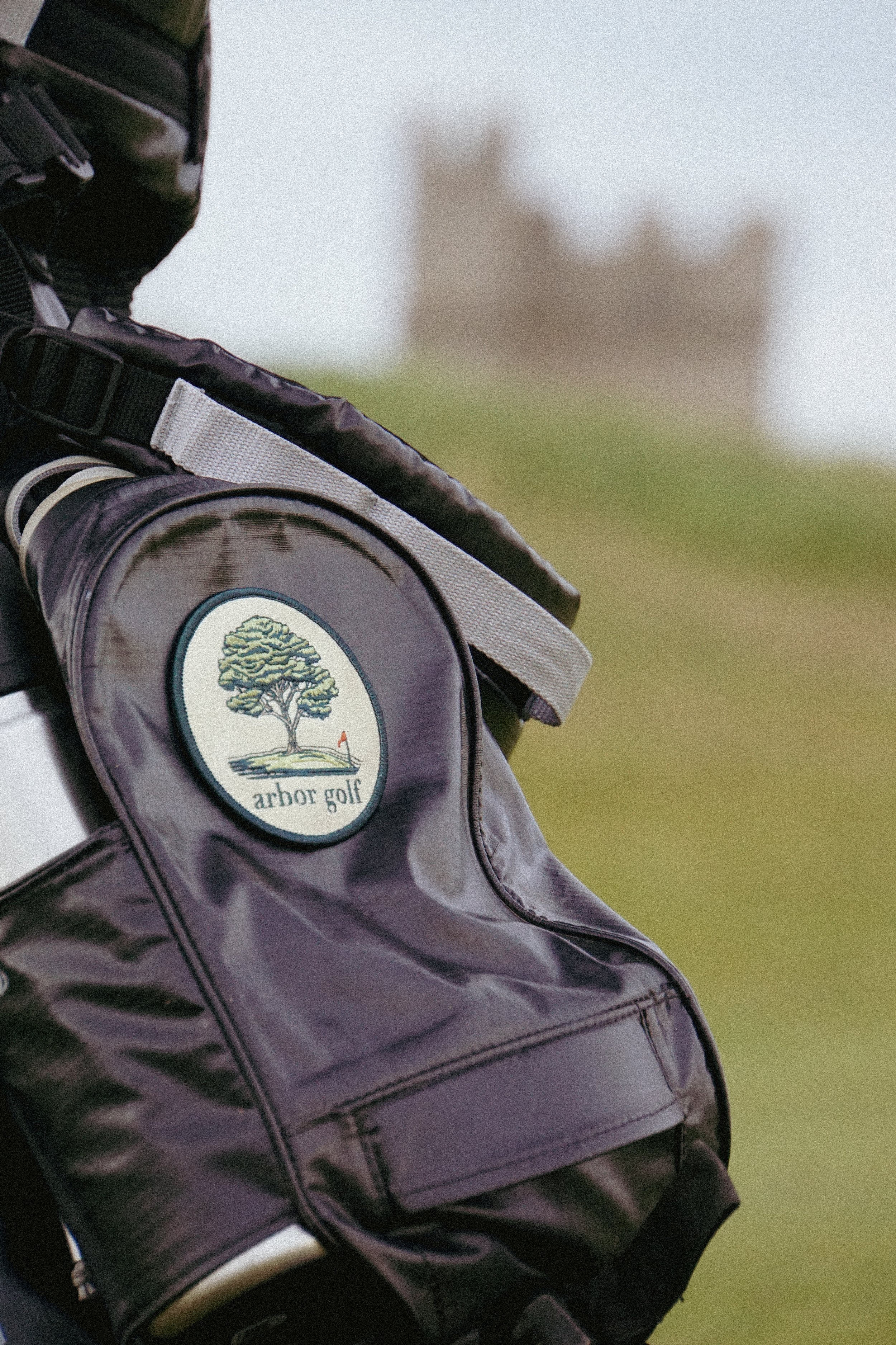 Close-up of a golf bag with "arbor golf" logo, featuring a tree and golf flag, with blurred scenic landscape in the background.