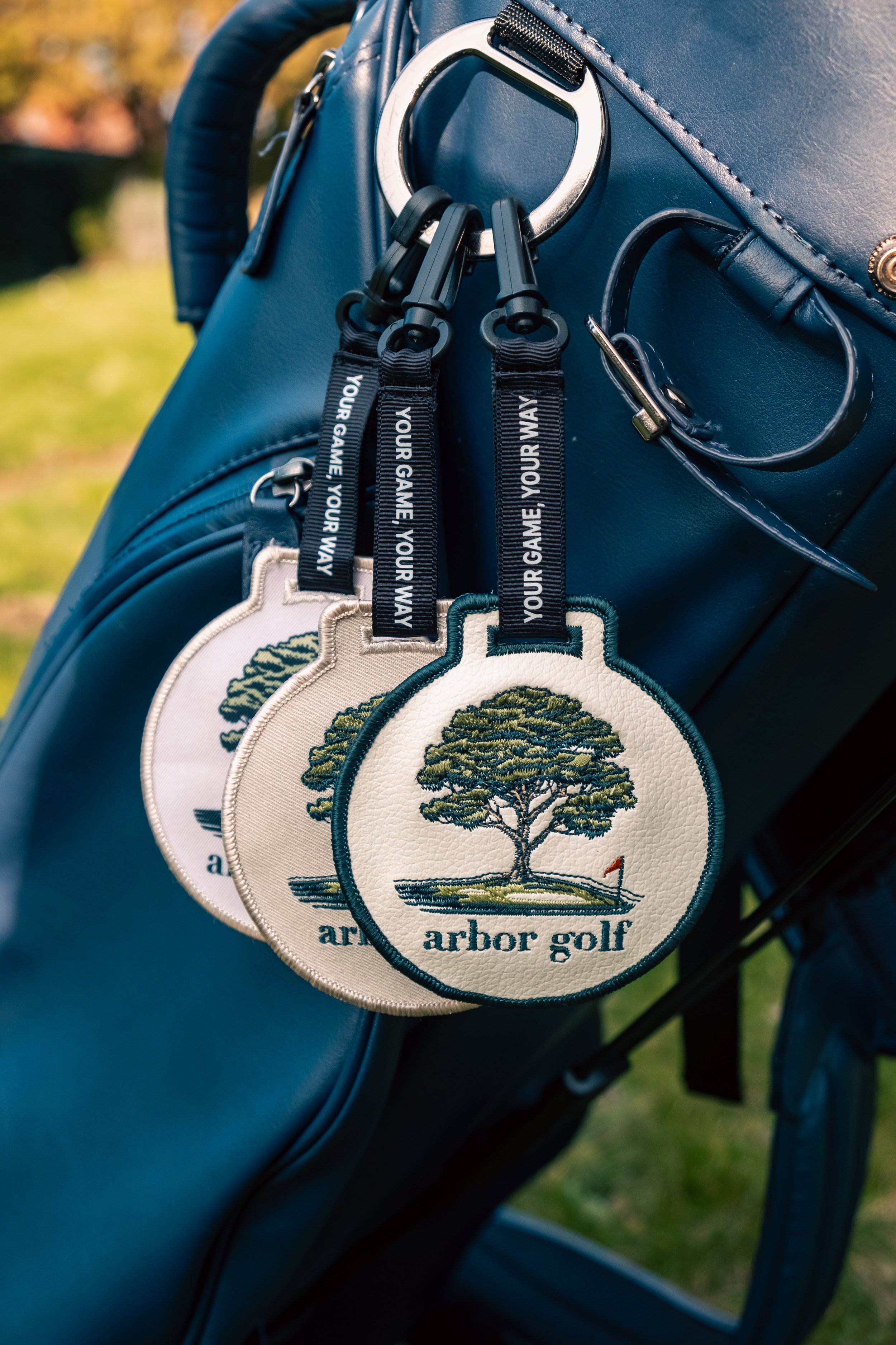 Close-up of a blue golf bag with three embroidered Arbor Golf tags, each showing a tree and flag design, attached with black straps featuring the text 'YOUR GAME, YOUR WAY'.