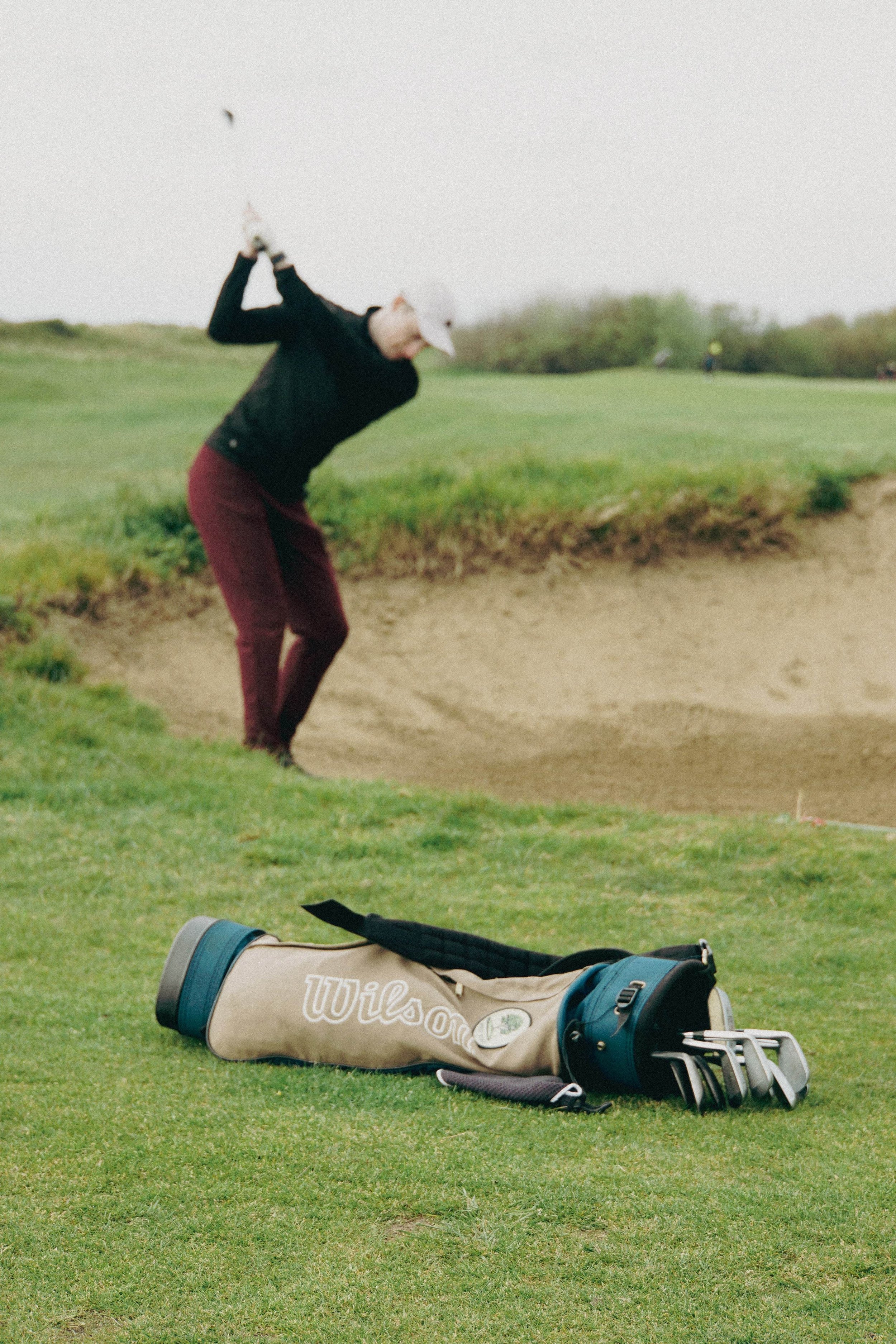 Golfer swinging near sand trap with golf bag and clubs on grass