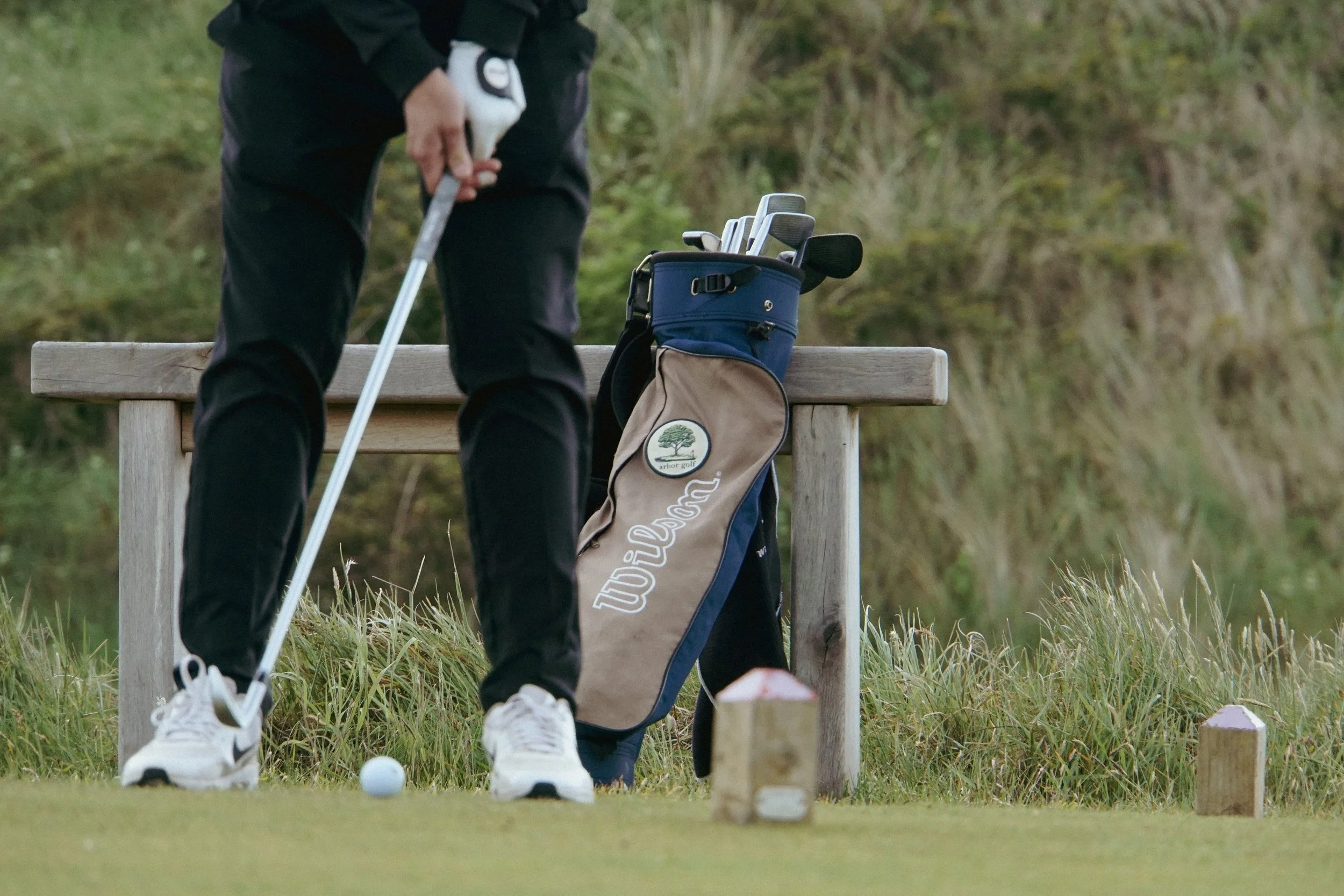 Golfer preparing to putt on a green, with a golf bag and clubs resting on a wooden bench nearby.