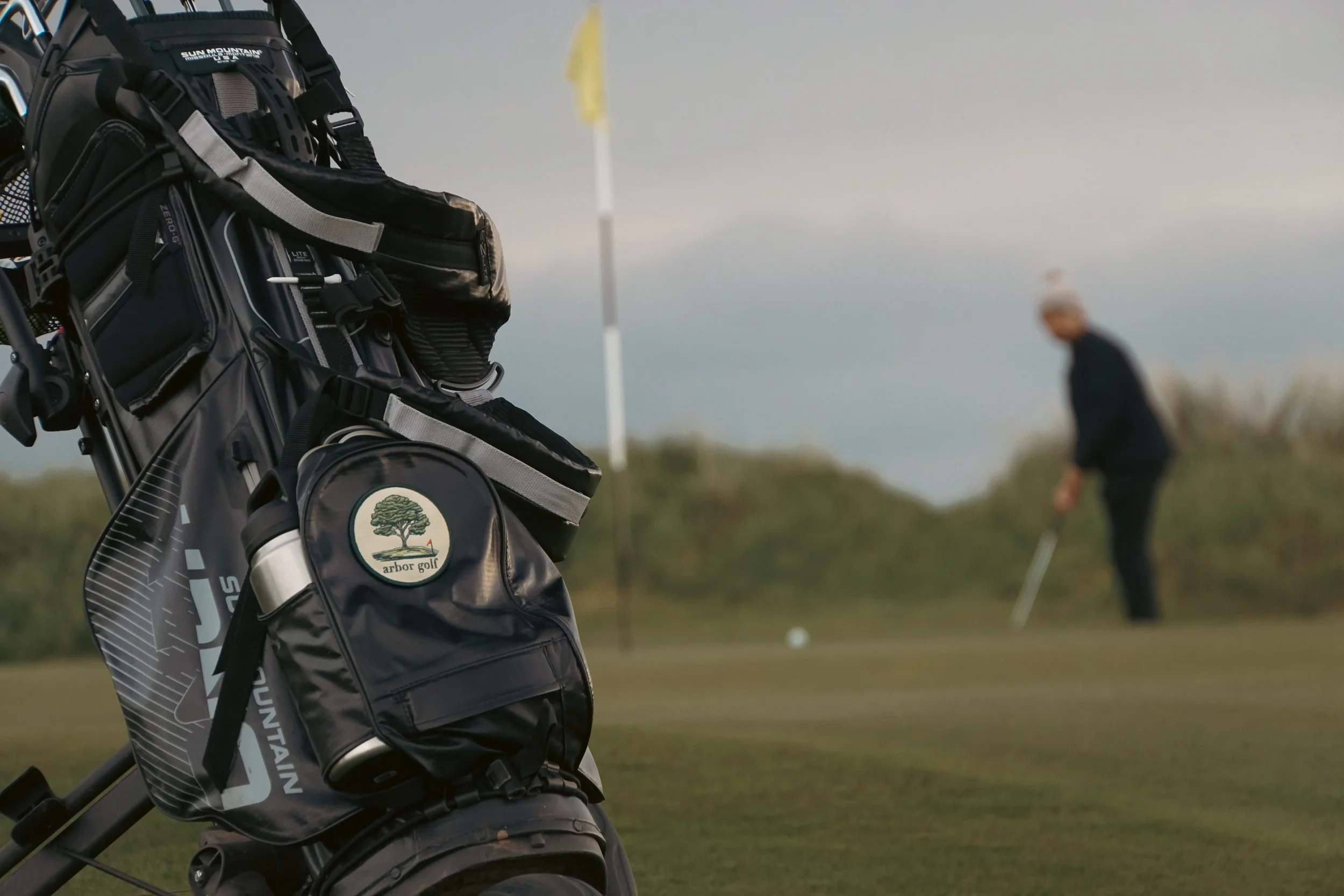 Golf bag with clubs and a logo that says 'arbor golf' in focus; golfer putting on the green in the background.