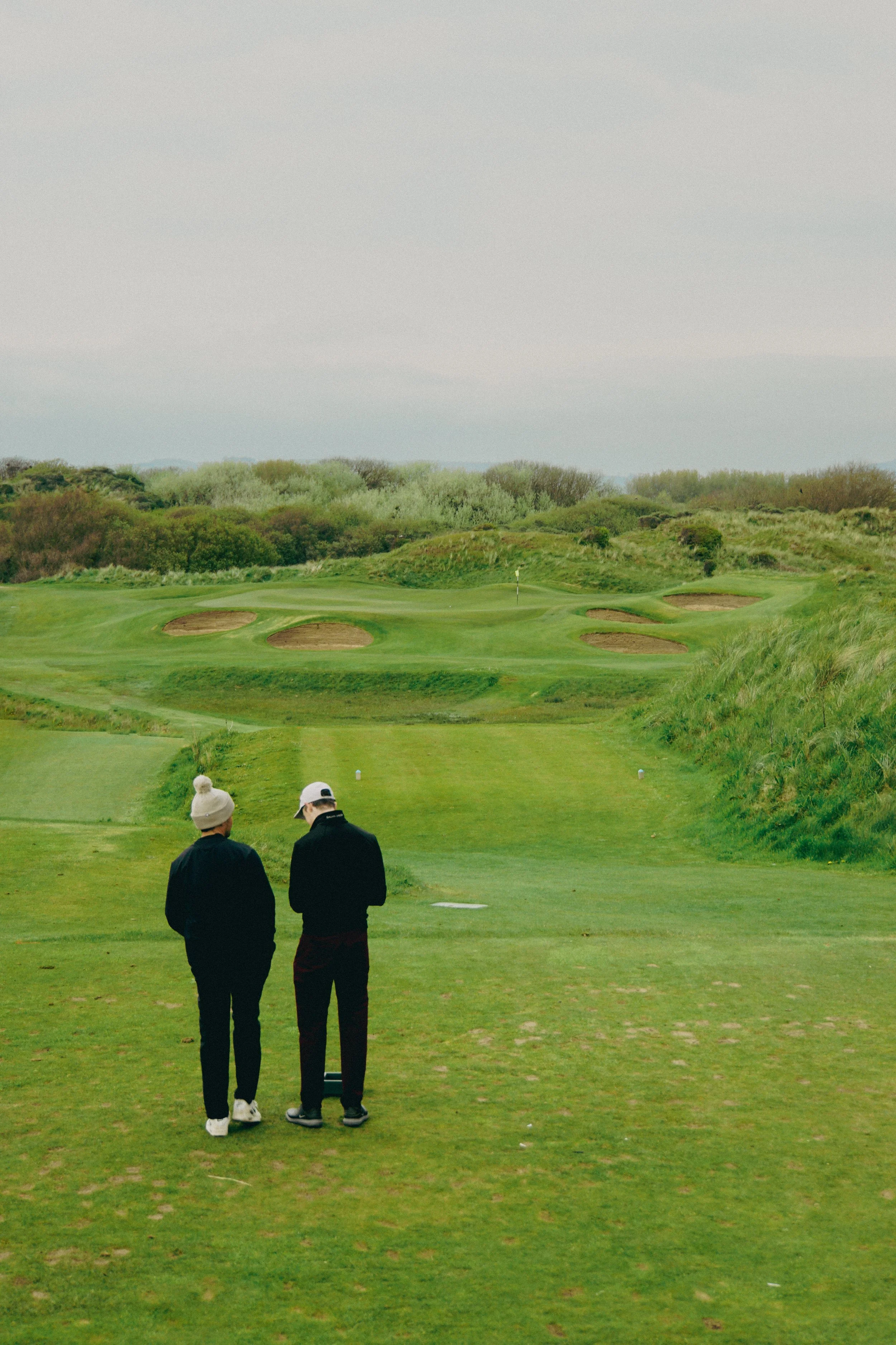 Two people standing on a golf course, looking toward a green with sand traps, surrounded by greenery.