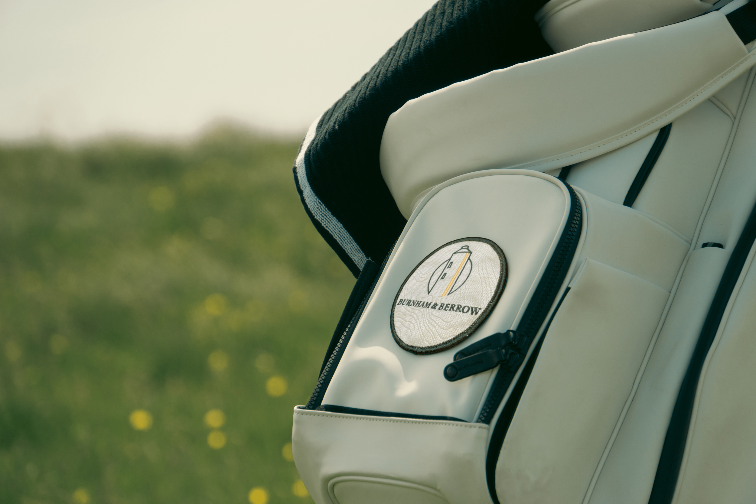 Close-up of a white golf bag with "Burnham & Berrow" logo, featuring a lighthouse, set against a grassy field background.